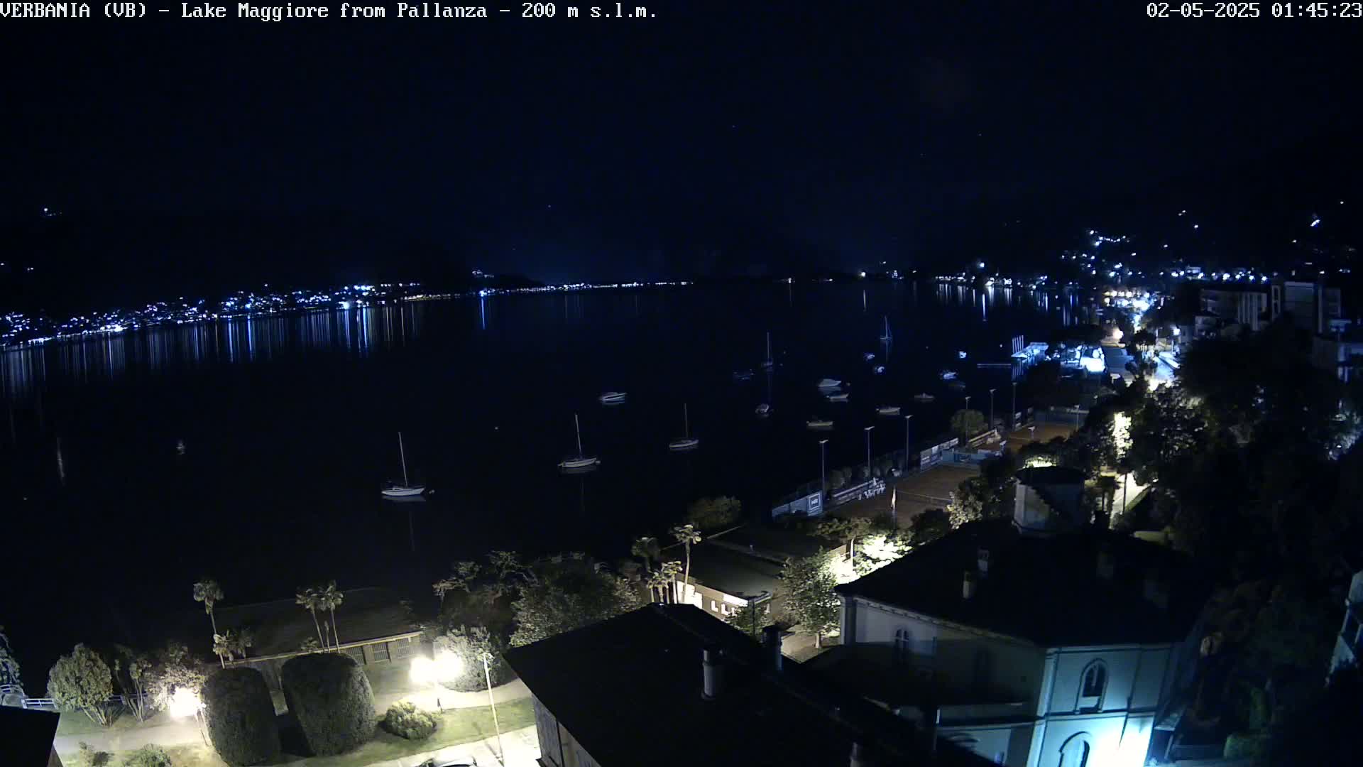 A nighttime view of a calm lake with several boats, a town with lights reflecting on the water, and a clear sky with stars.