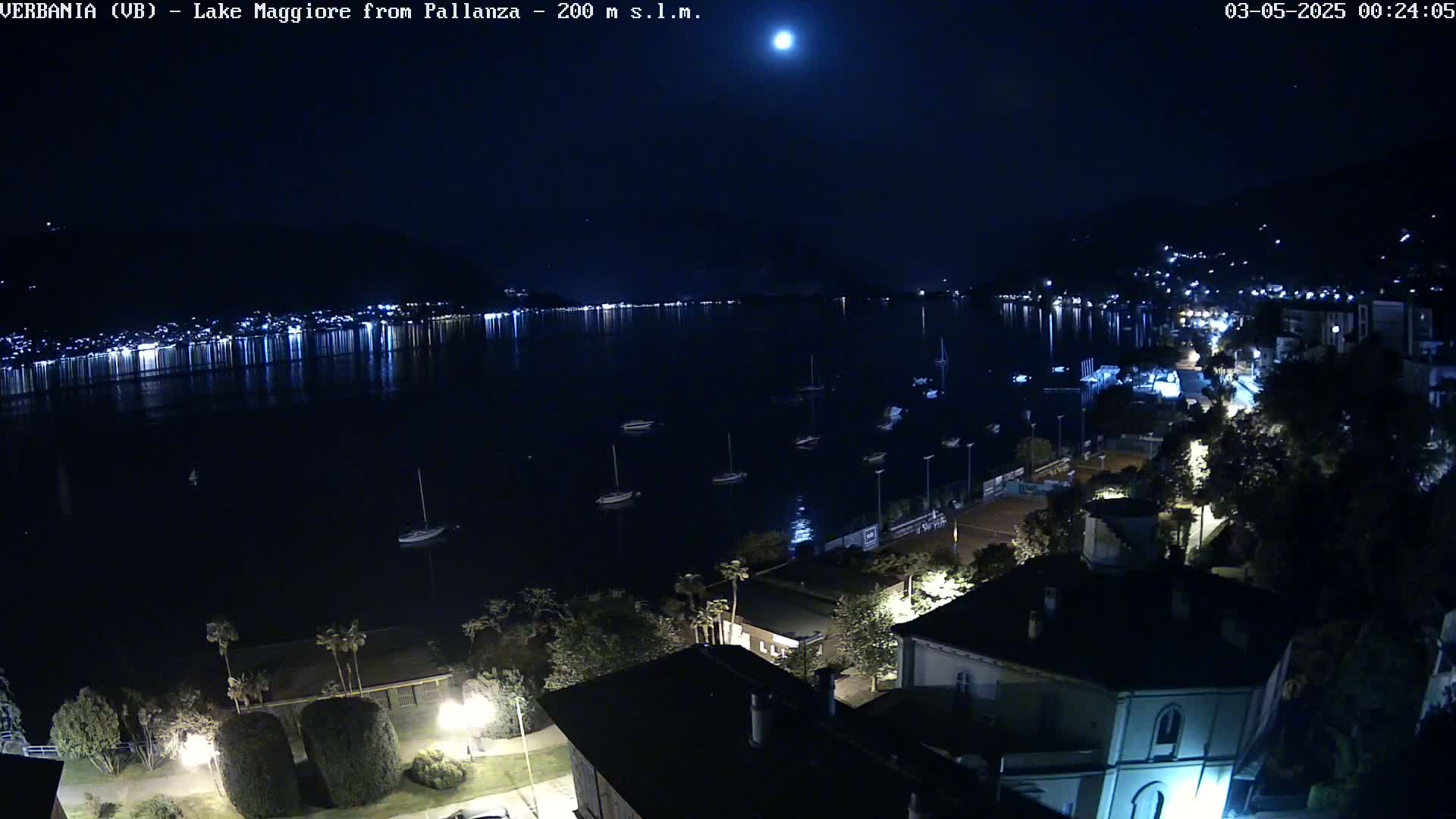 A nighttime view of a moonlit lake with several boats moored, a town's lights reflected on the water, and buildings illuminated along a shoreline.