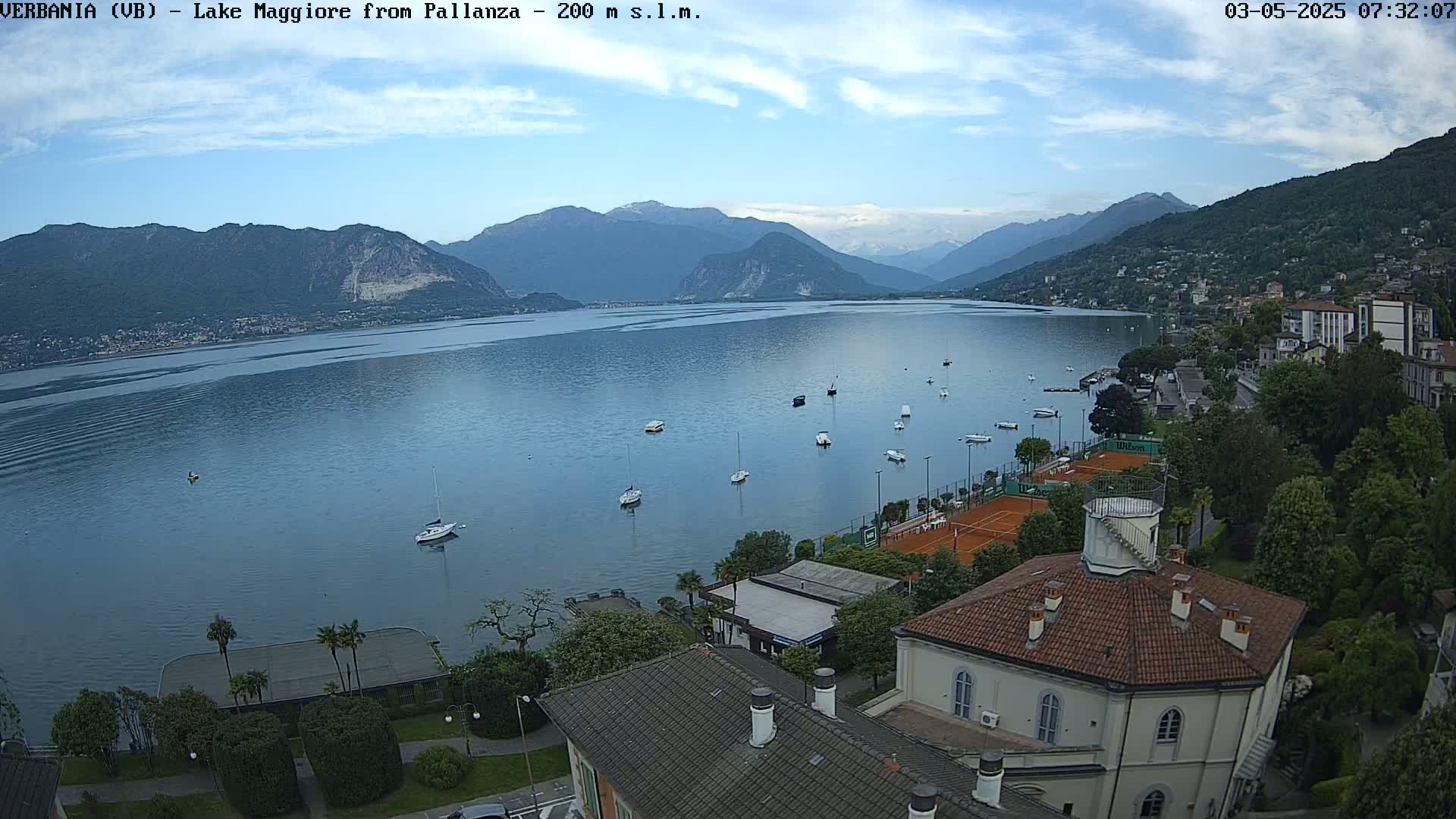 A calm lake with numerous boats is seen from an elevated vantage point, overlooking a town nestled against mountains under a partly cloudy sky.