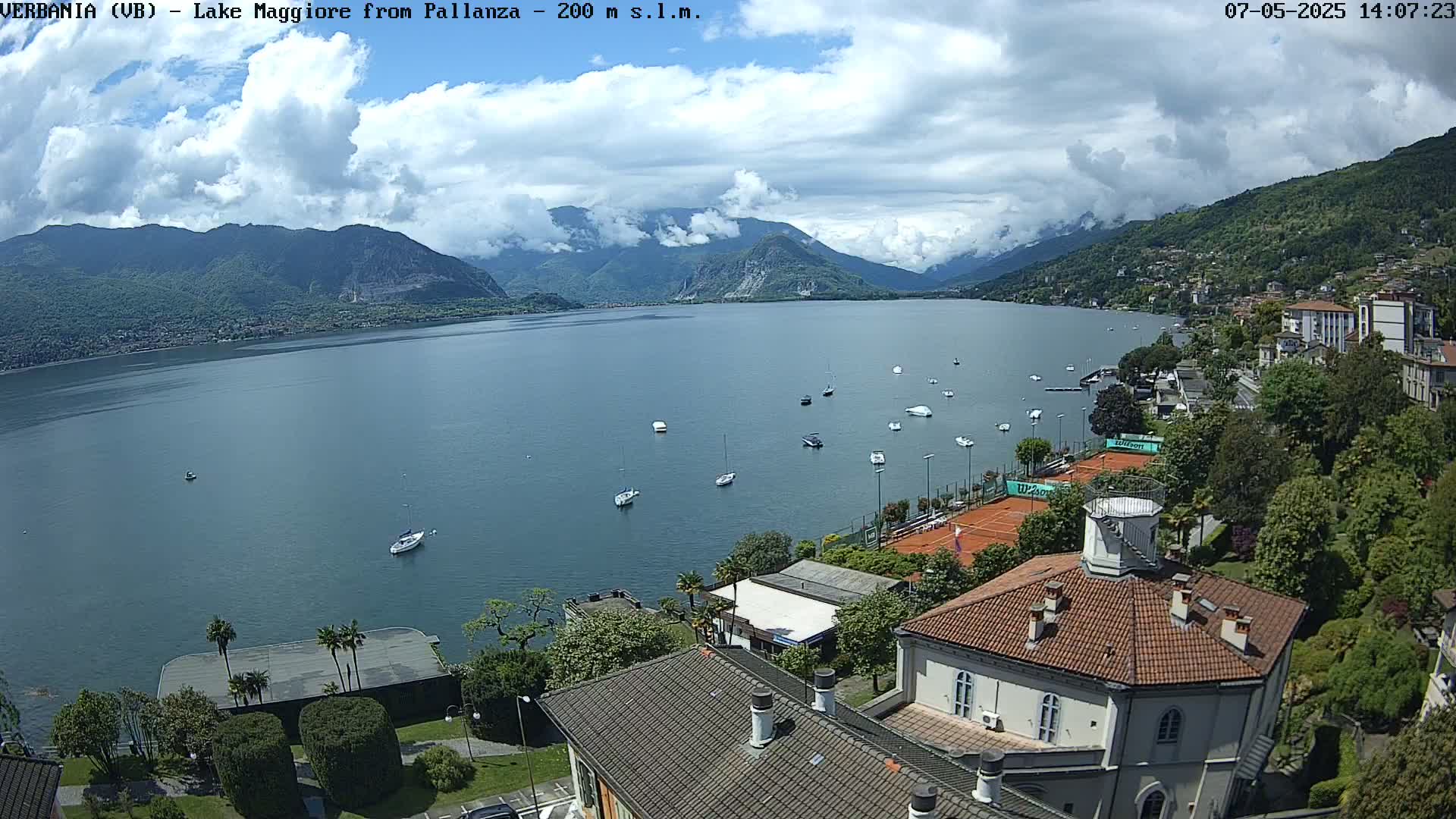 A partly cloudy day overlooking a calm lake with numerous boats, tennis courts, and buildings nestled against a mountainside.