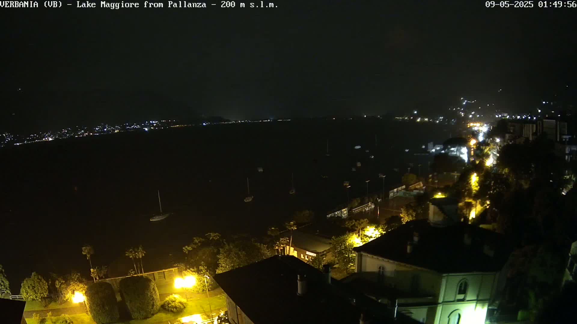 A nighttime, high-angle view shows a dark lake with sparsely scattered boats and a town's lights illuminating buildings and streets along the shoreline.