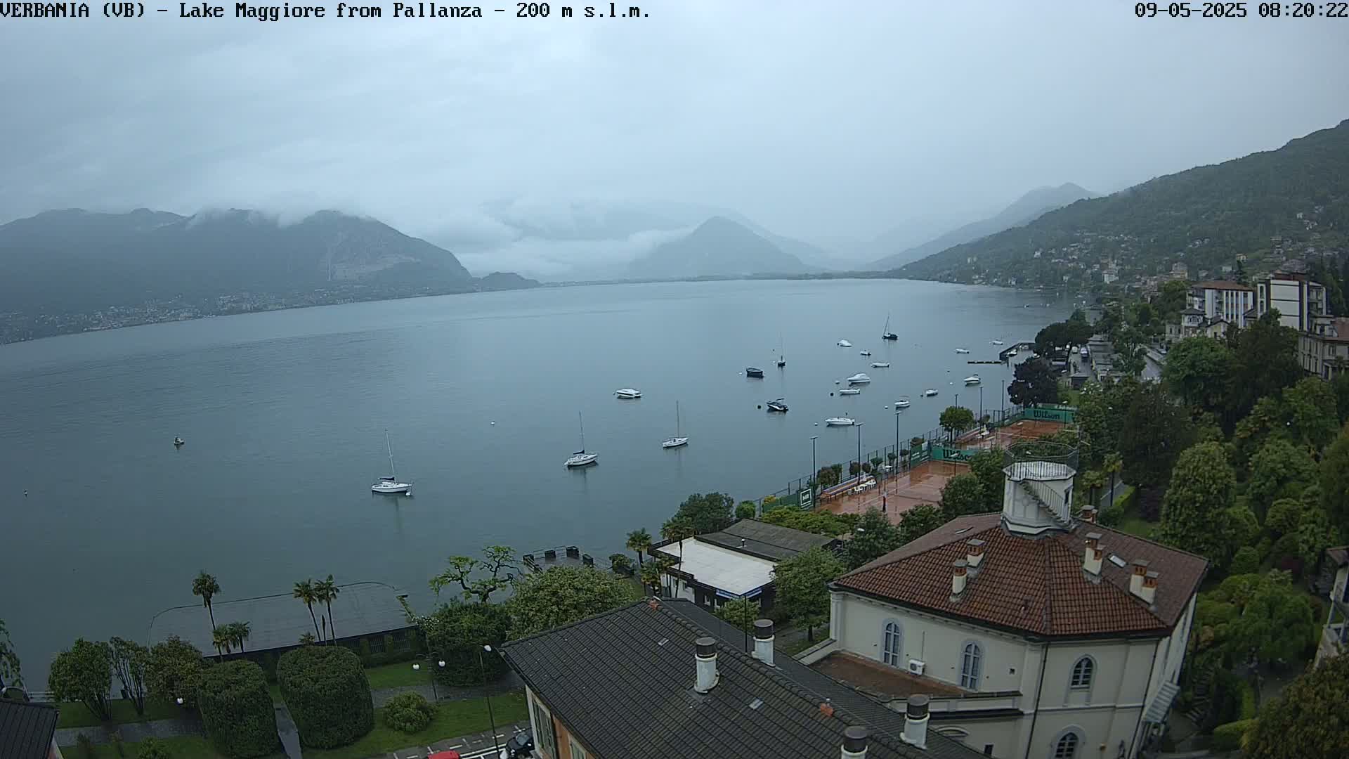 A calm lake surrounded by mountains and buildings under an overcast sky, with several boats and a tennis court visible.