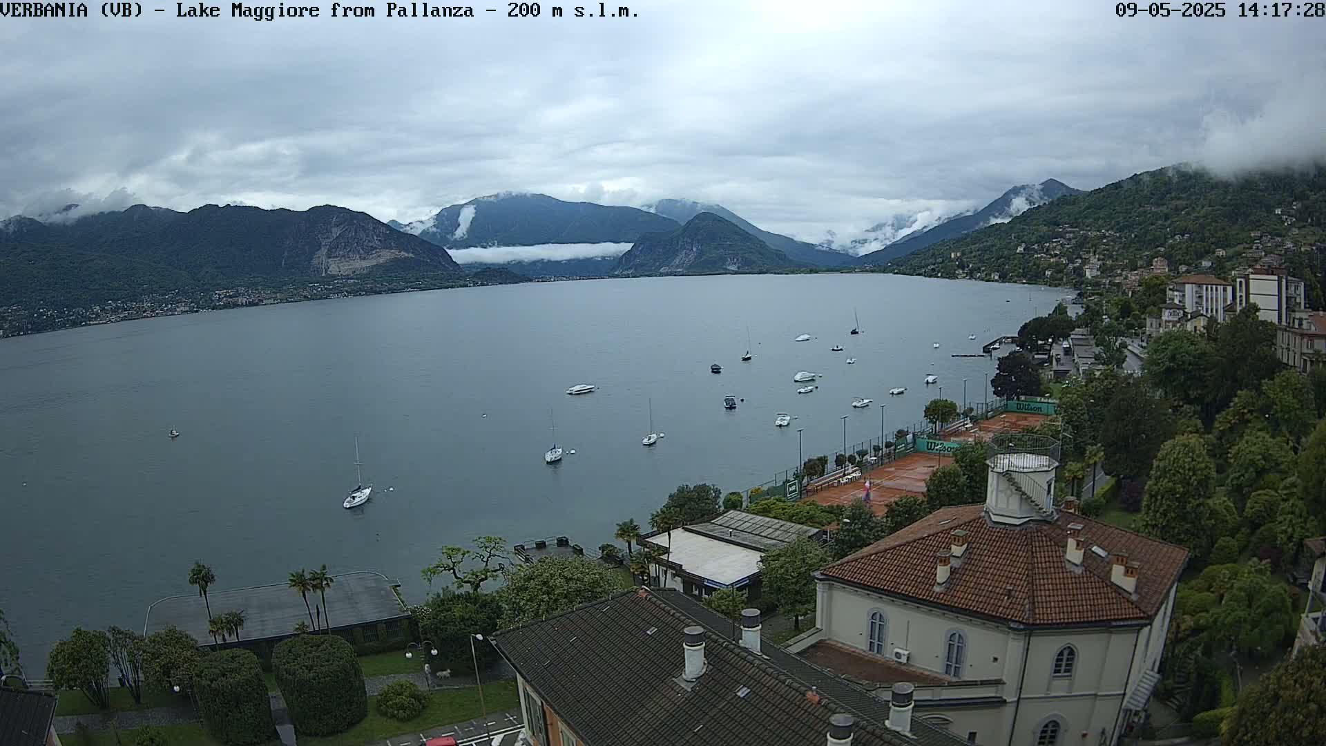 An overcast day reveals a calm lake with several sailboats, a shoreline town, and mountains in the distance.