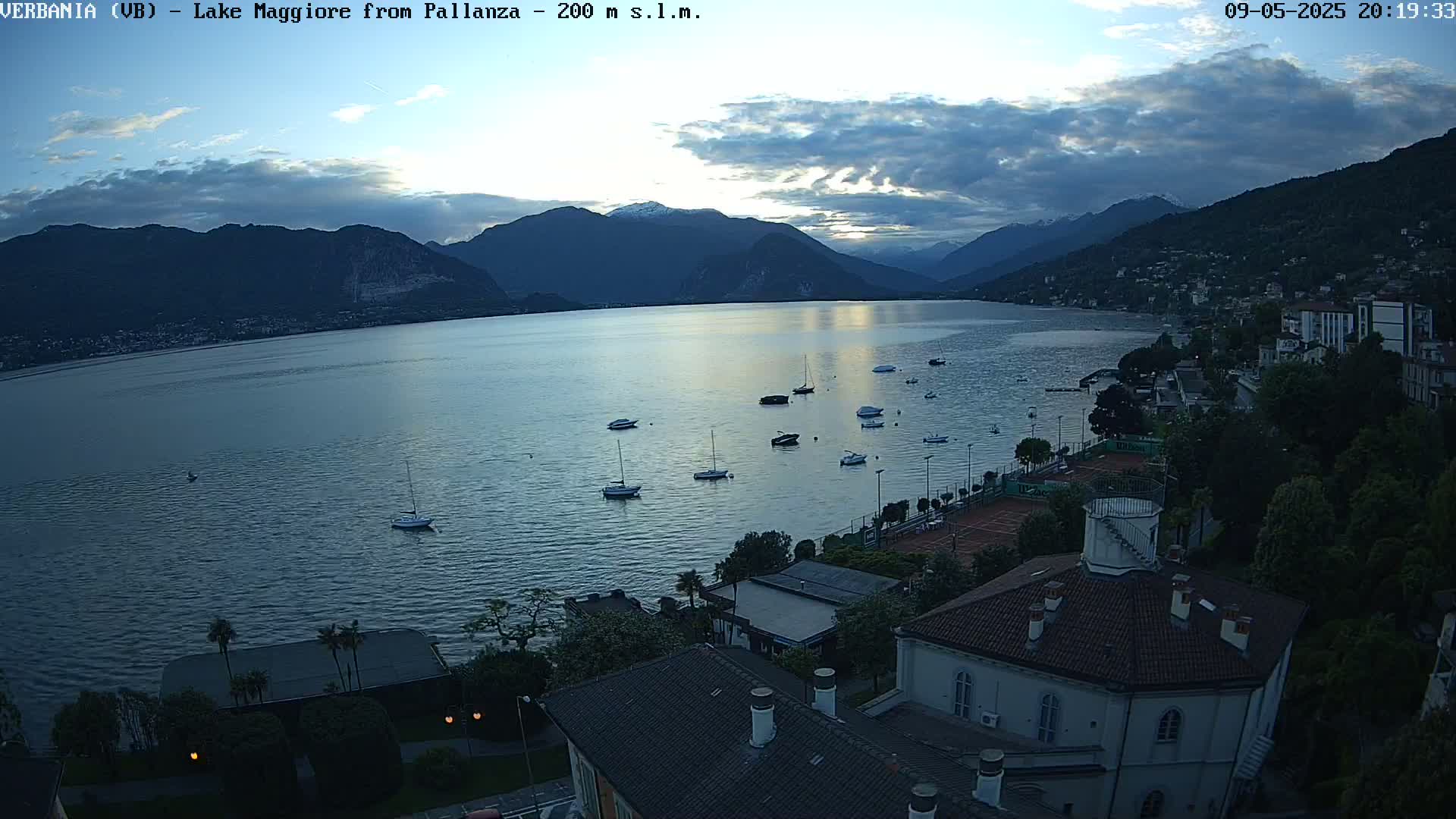 An elevated view shows a calm lake with several boats at dusk, under a partly cloudy sky, with a town and mountains in the background.