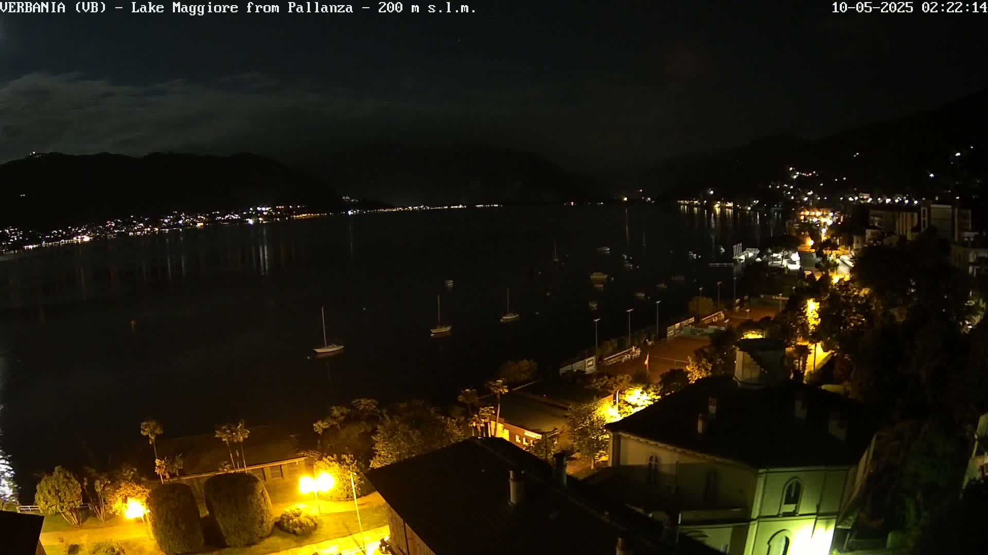 A nighttime view of a calm lake with several small boats moored, a town illuminated with streetlights along the shoreline, and dark mountains in the background under a clear night sky.