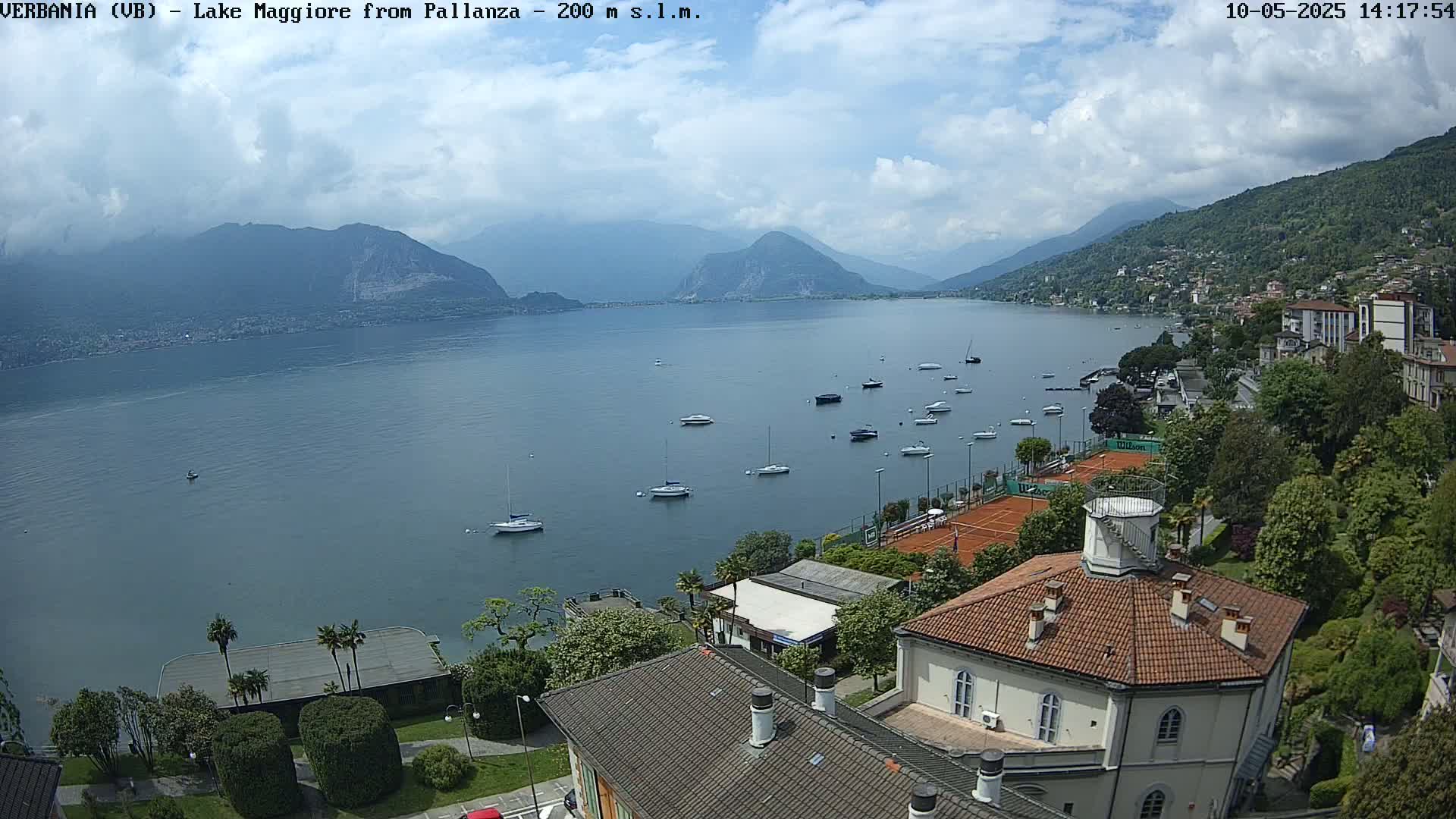 A partly cloudy day overlooking a calm lake with numerous boats and a tennis court, nestled among green vegetation and buildings.