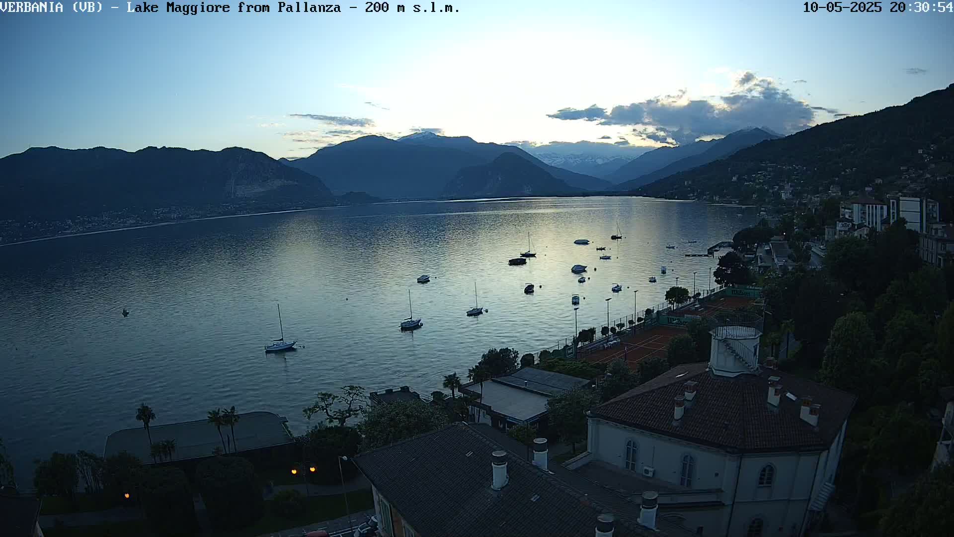 An elevated view shows a calm lake at dusk, surrounded by mountains and buildings, with several boats on the water and tennis courts visible near the shoreline.
