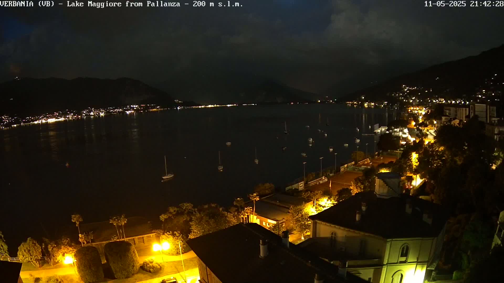 A nighttime view of a calm lake with boats and a city illuminated by lights on a hill under a partly cloudy sky.