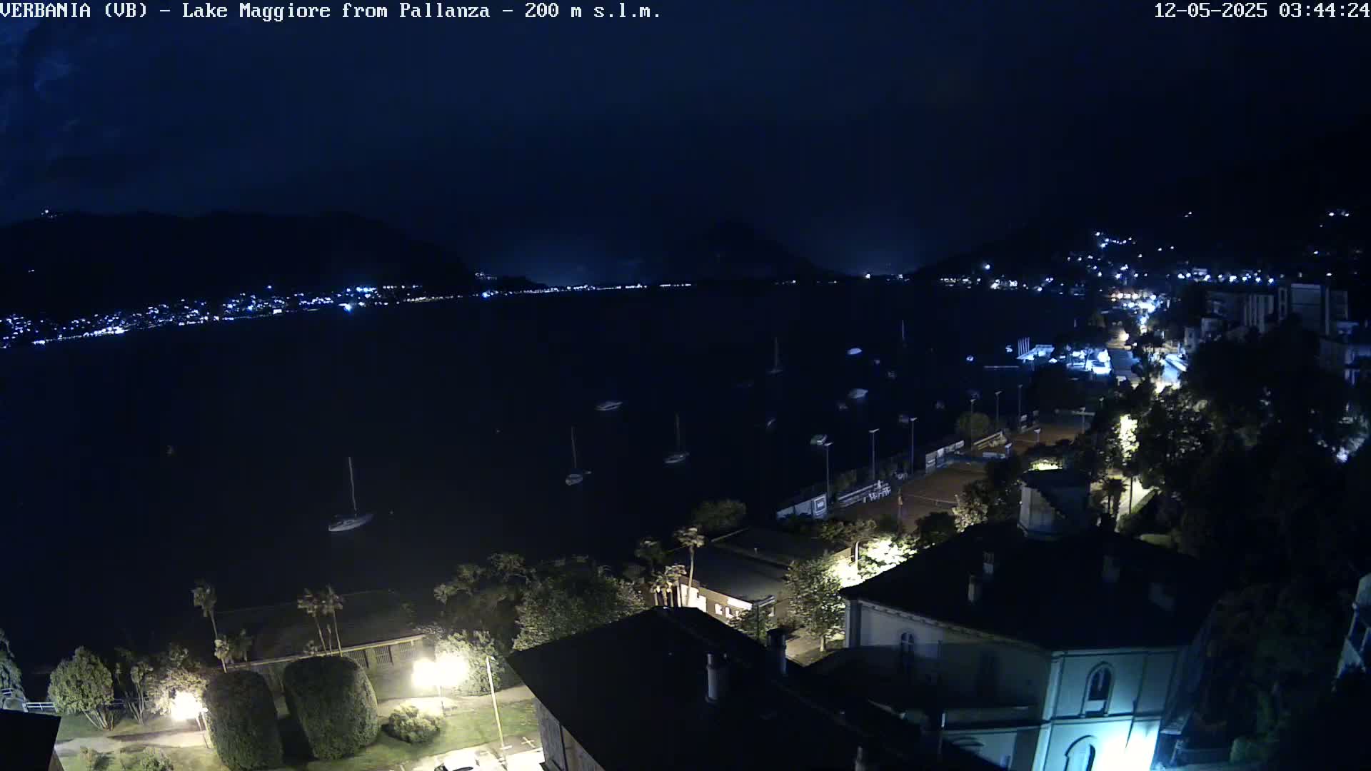 A nighttime, high-angle view shows a dark lake with several boats, a shoreline with buildings and streetlights, and hills in the background under a clear, dark sky.