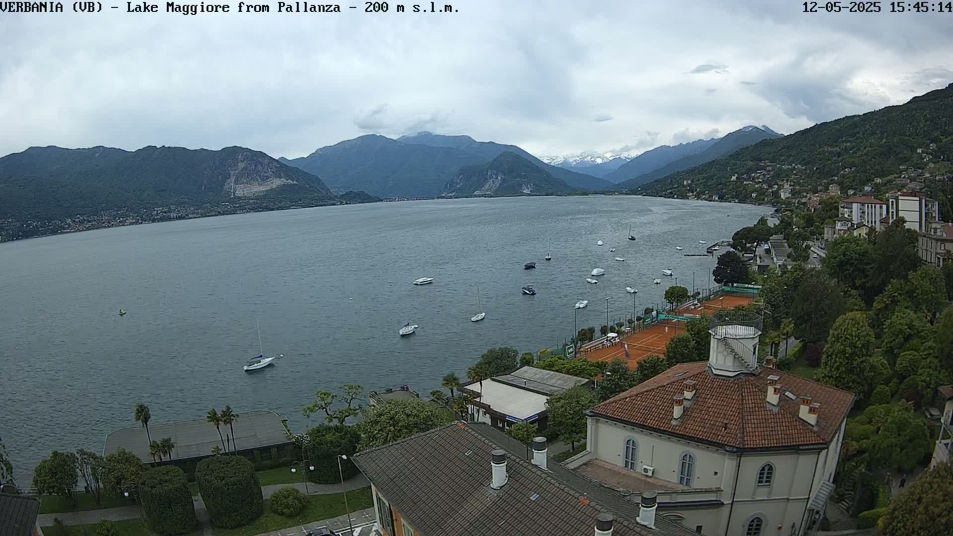 A view of Lake Maggiore from a hilltop, under a partly cloudy sky, shows several boats on the water, tennis courts, and buildings along the shoreline.