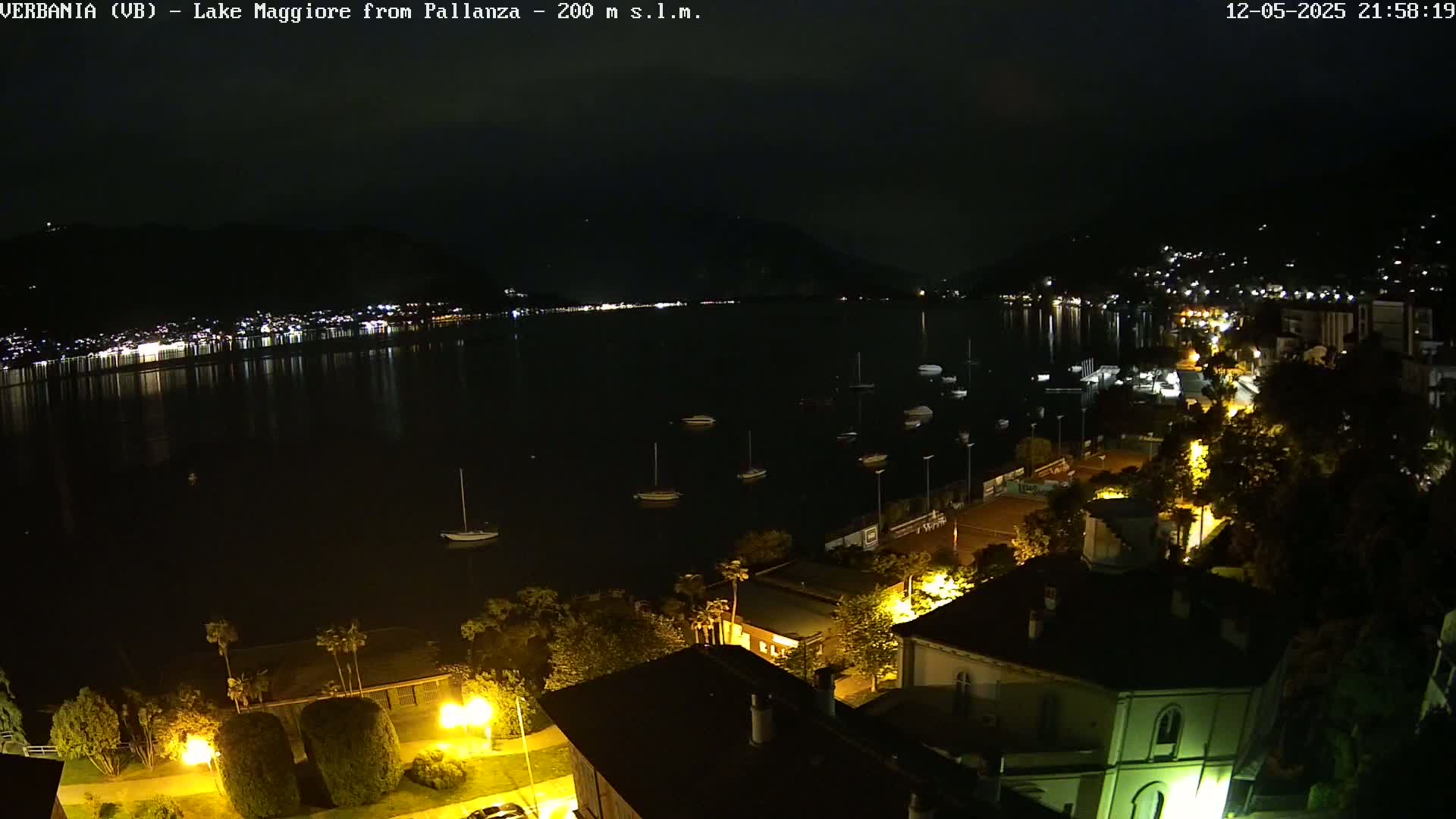 A nighttime view of a lake with boats, illuminated buildings along the shoreline, and dark hills in the background under a clear sky.