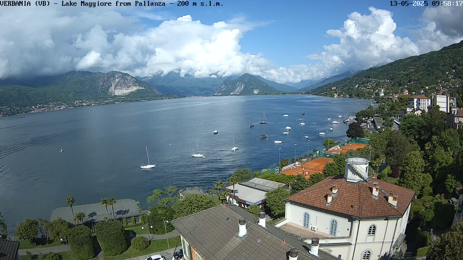 A partly cloudy day overlooks a calm lake with several boats, tennis courts, and buildings nestled along the shoreline, backed by mountains.