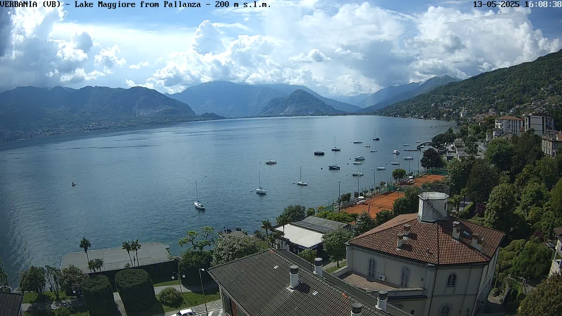 A partly cloudy day reveals a tranquil lake with numerous boats, surrounded by lush greenery, mountains, and buildings, including a round-roofed structure.