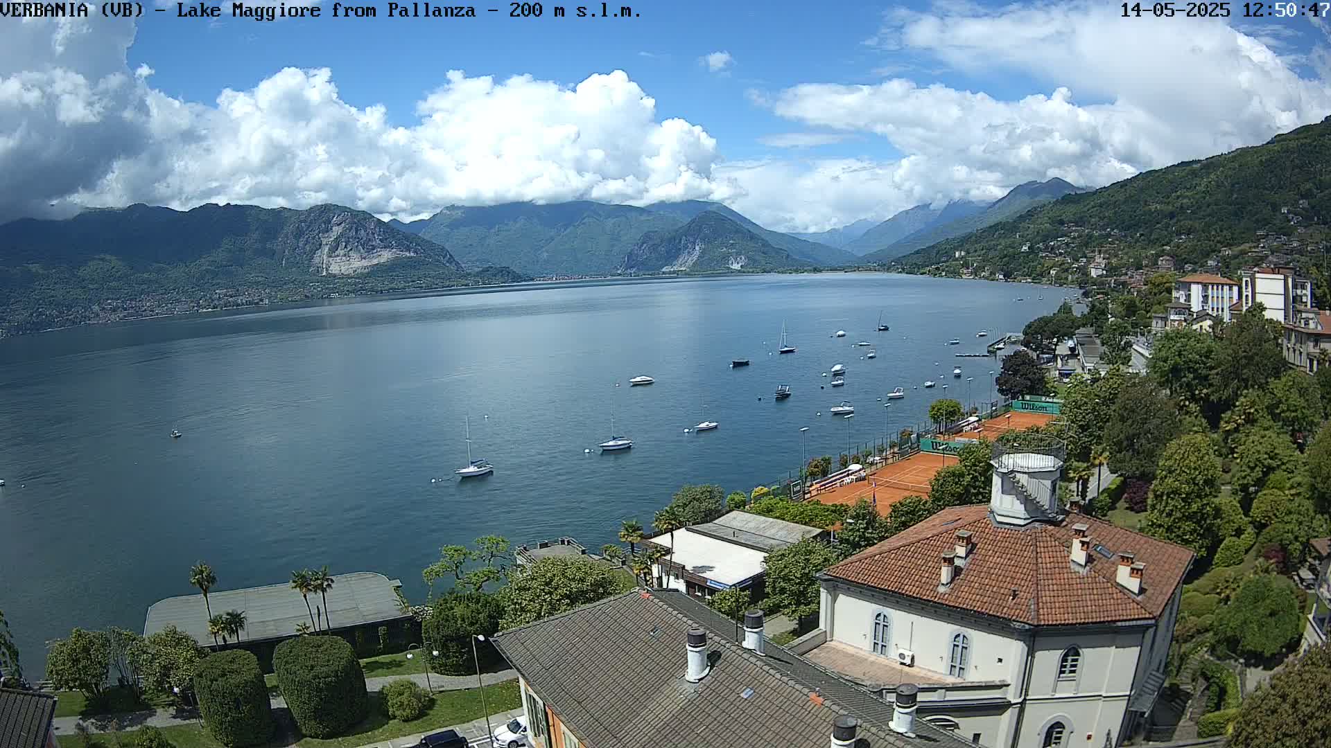 A sunny day overlooking a calm lake dotted with sailboats, surrounded by mountains and a lakeside town with tennis courts and buildings.