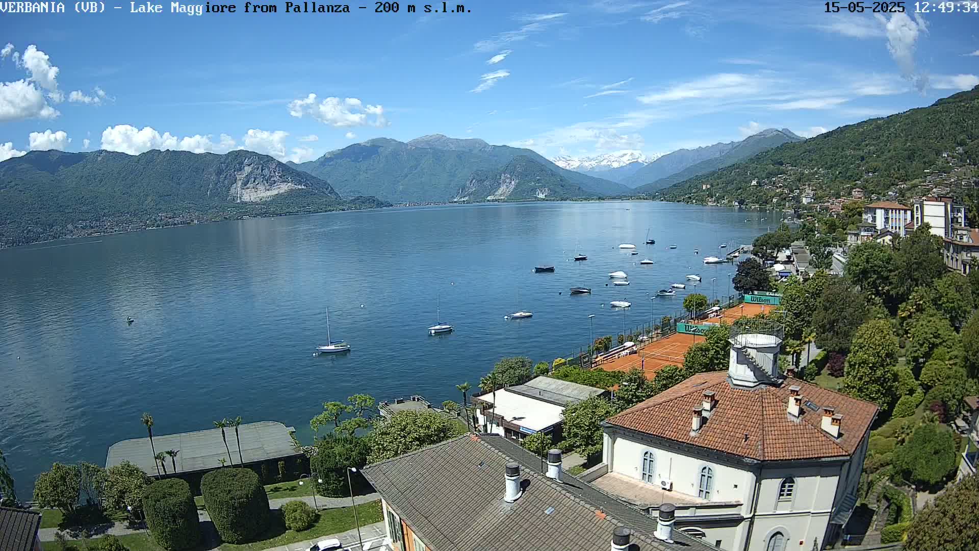 A sunny day overlooking a calm lake with numerous boats, tennis courts, and buildings nestled against a backdrop of mountains.