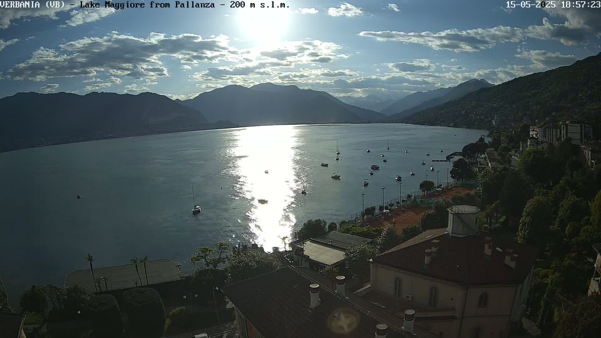 A partly cloudy afternoon shows a tranquil lake with several boats, surrounded by mountains and buildings with tennis courts.