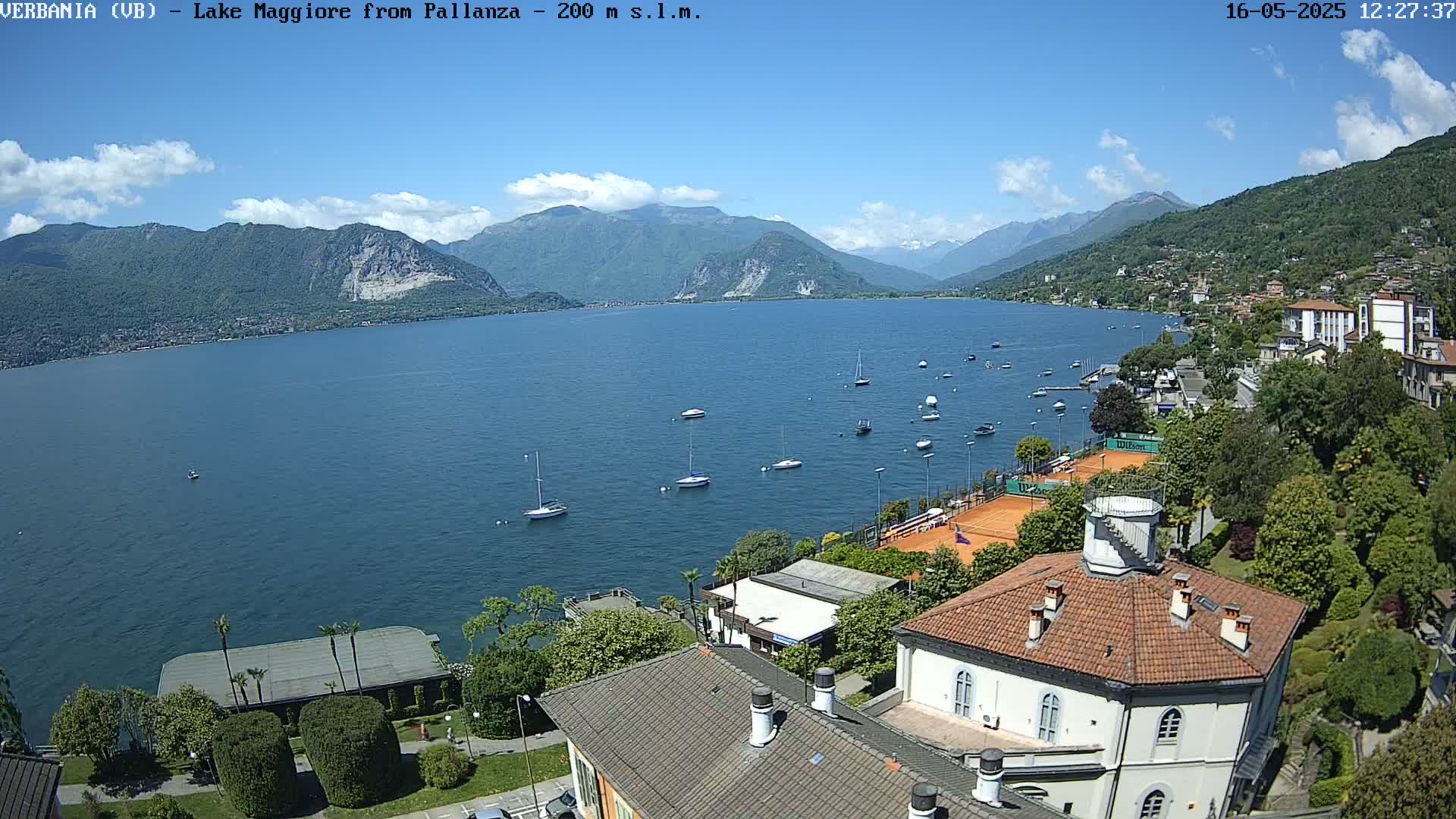 A clear, sunny day overlooking a calm lake with several boats, tennis courts, and buildings nestled into a hillside.