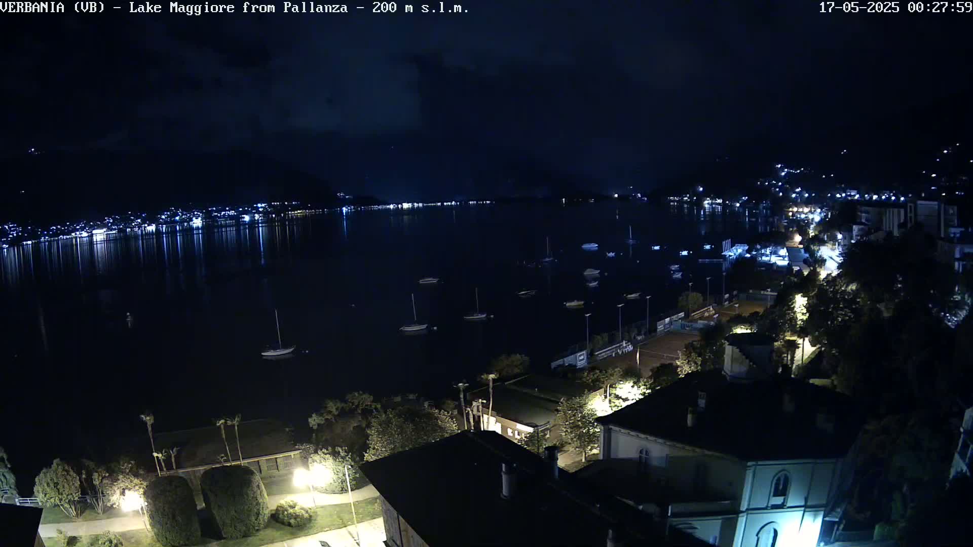 A nighttime, high-angle view shows a calm lake reflecting the lights of a town on its shore, with several small boats moored in the water and buildings, trees, and streetlights visible on land.