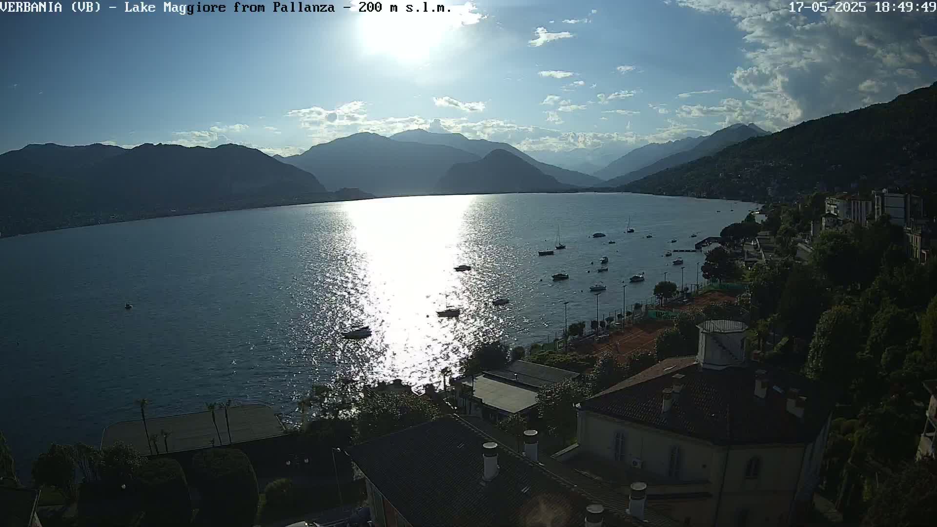 A partly sunny day shows a calm lake with several boats, surrounded by mountains and a shoreline with buildings and trees.