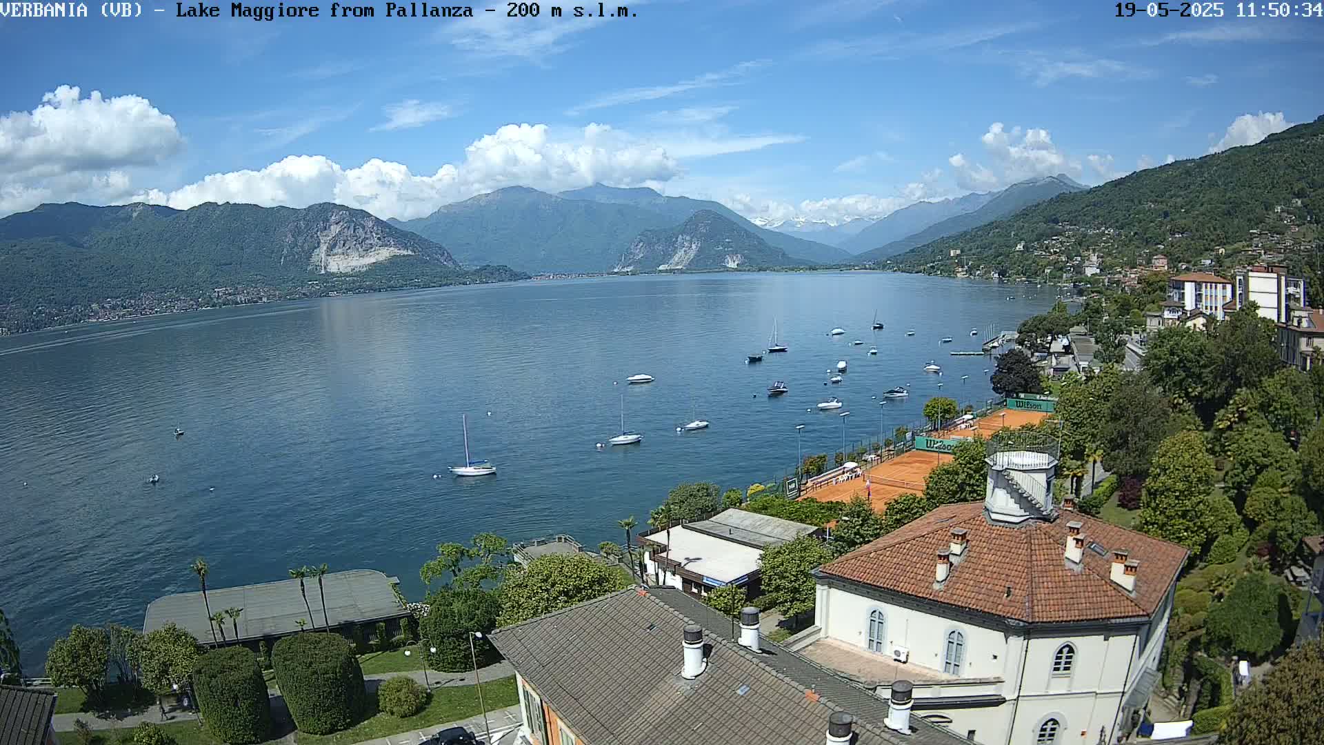 A partly cloudy day shows Lake Maggiore, numerous sailboats on the water, tennis courts, and buildings nestled against a hillside, with mountains in the background.