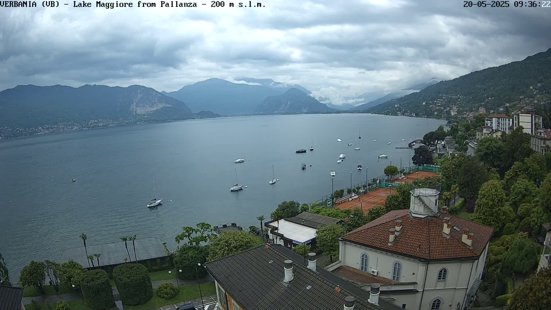A calm lake is seen under a cloudy sky, with several boats on the water and a town with buildings and tennis courts visible along the shoreline, nestled against mountains in the background.