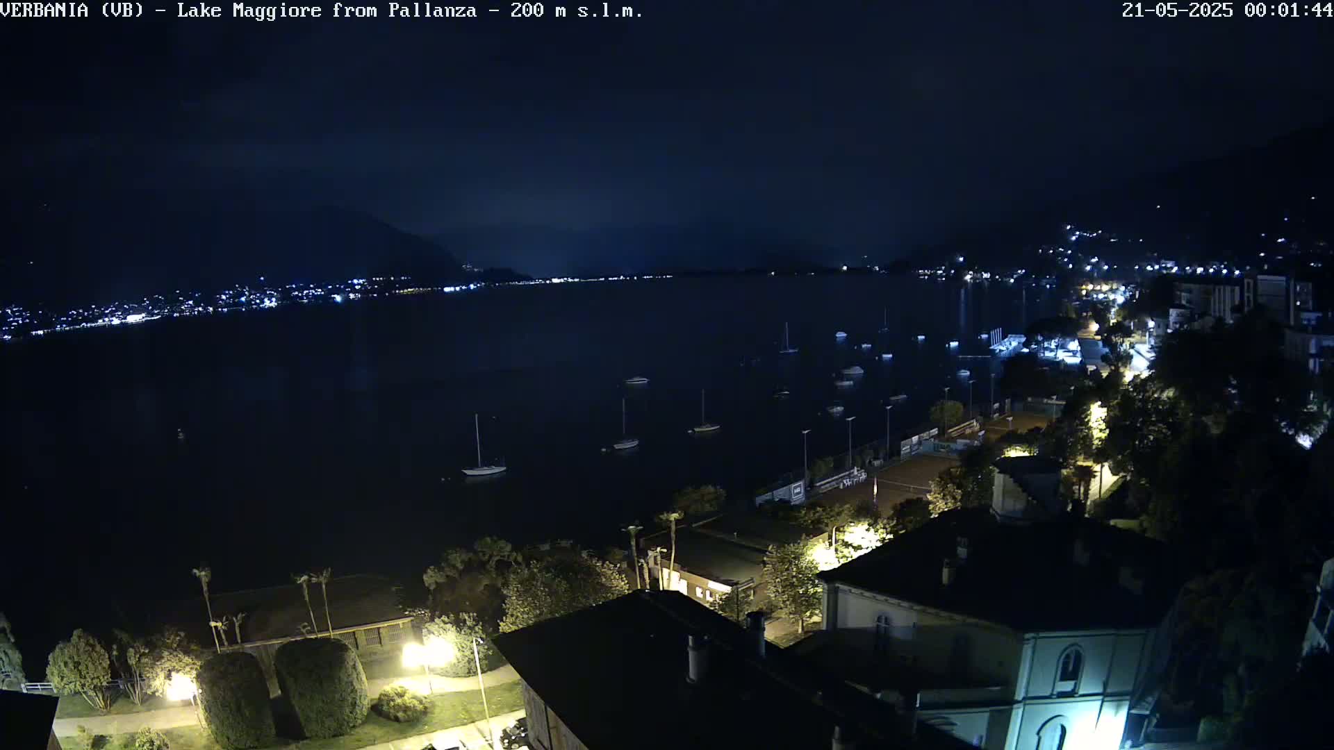 A nighttime, aerial view of a town situated on a lake, with boats moored on the water and lights illuminating the buildings and streets.