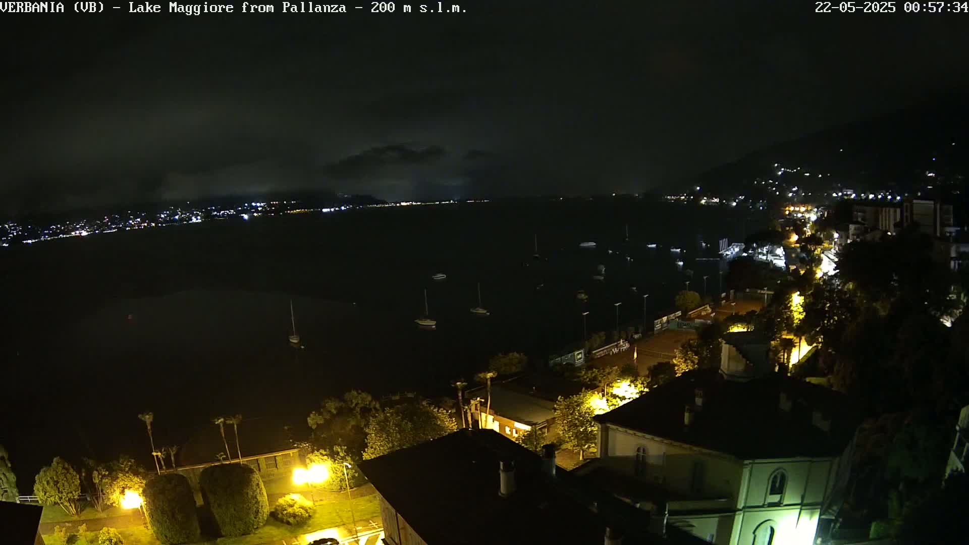 A nighttime view of a town situated on a lake, with several boats visible on the water under a partly cloudy sky.