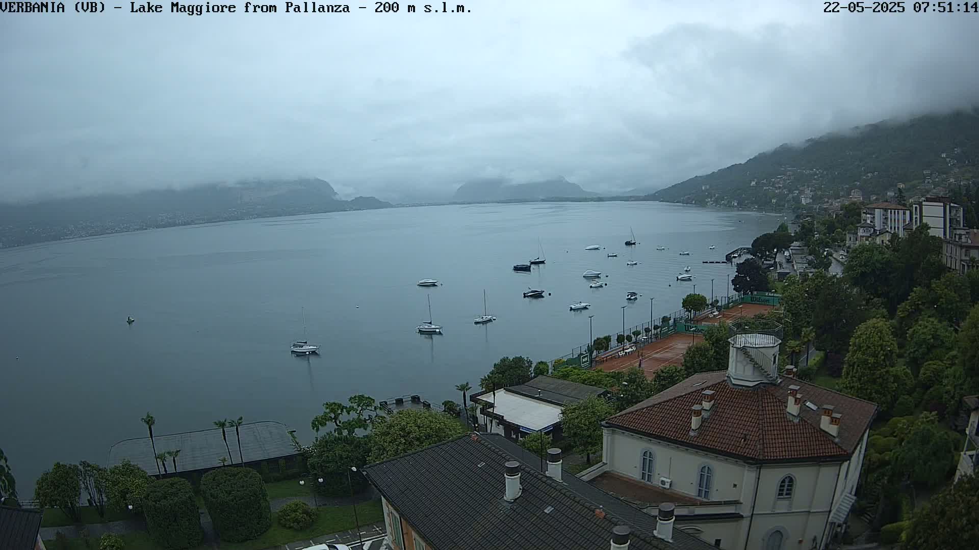 A cloudy day overlooks a calm lake with several boats, a tennis court, and buildings nestled along the shoreline.