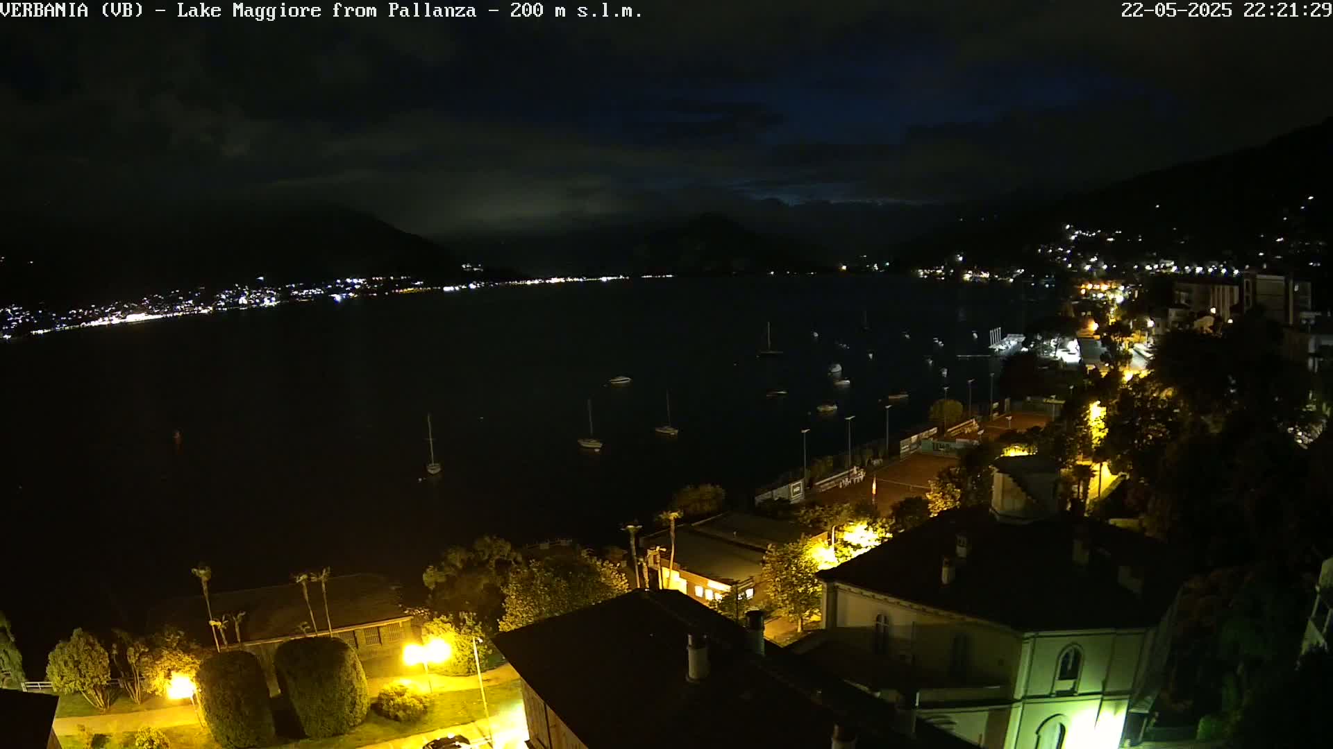 A nighttime view of a lake with boats, a town illuminated by streetlights, and dark hills in the background under a mostly cloudy sky.