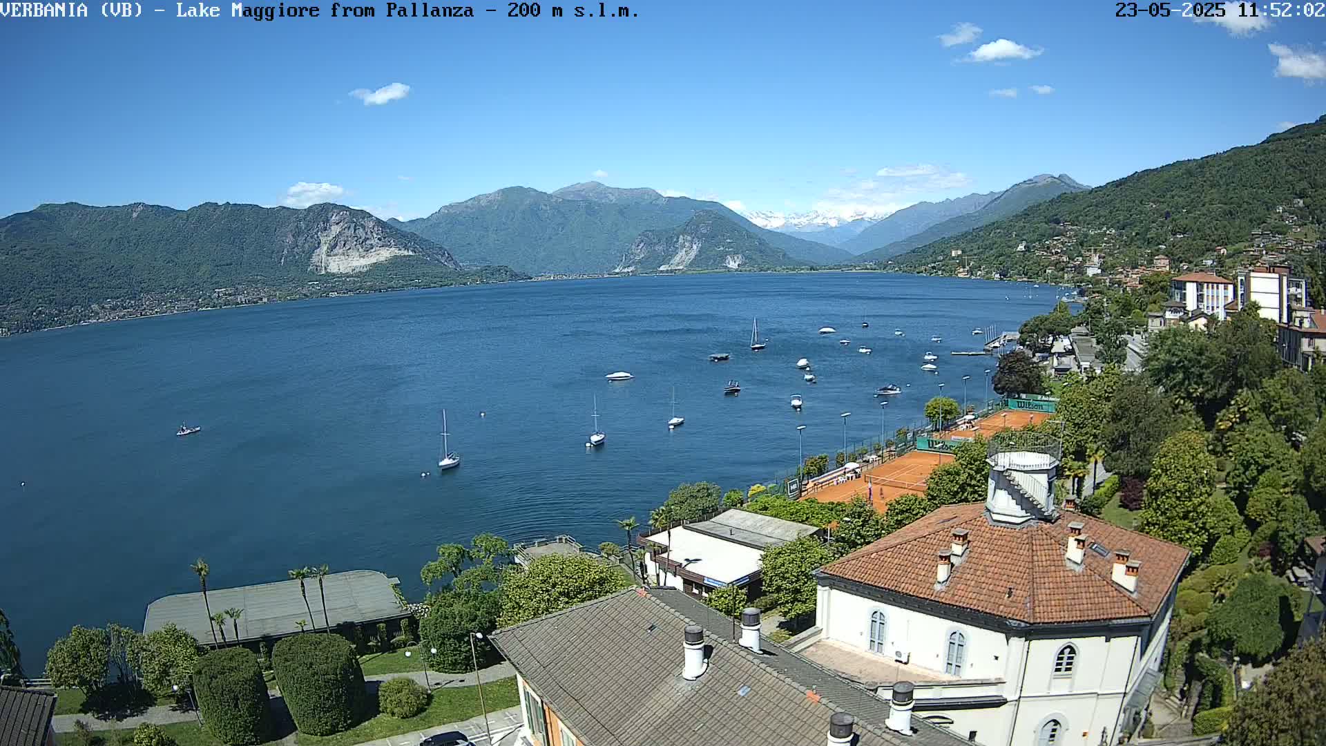 A sunny day overlooking a lake with numerous boats, tennis courts, and buildings nestled along the shoreline, backed by mountains.