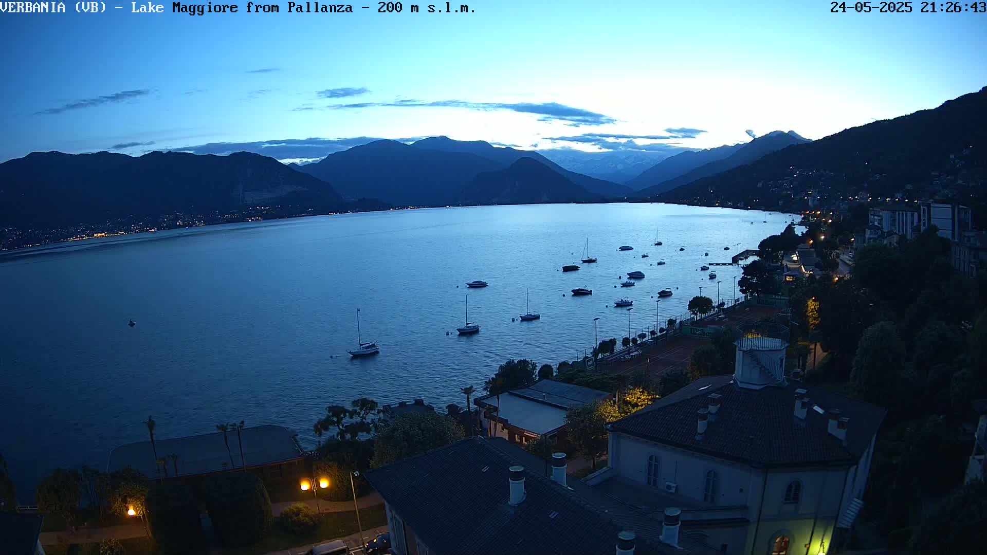 A twilight view of a lake with numerous boats, surrounded by dark mountains and a shoreline with buildings and lit tennis courts.
