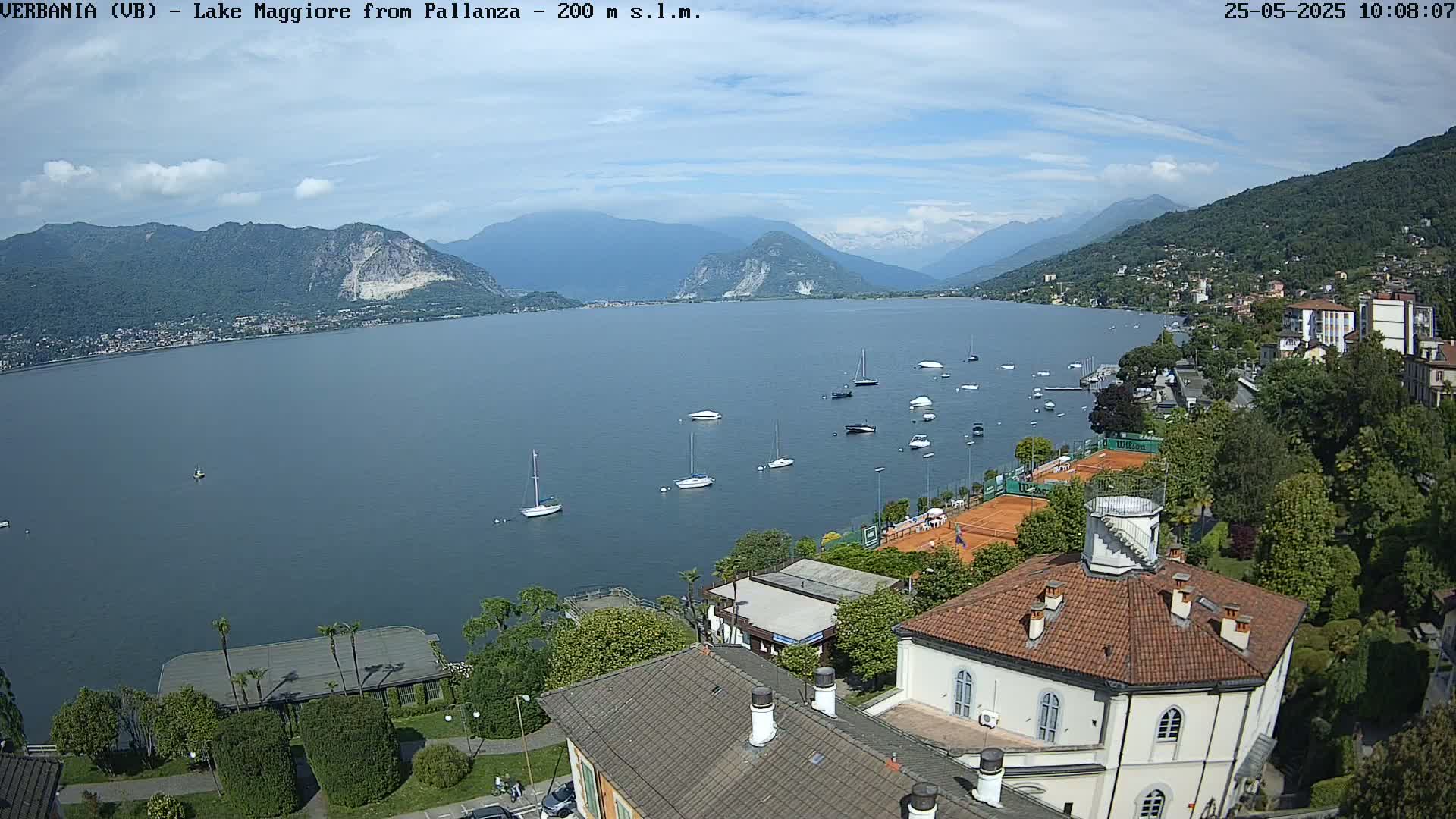 A sunny day overlooking Lake Maggiore, with several sailboats on the water, a tennis court, and buildings nestled along the shoreline, mountains in the distance.