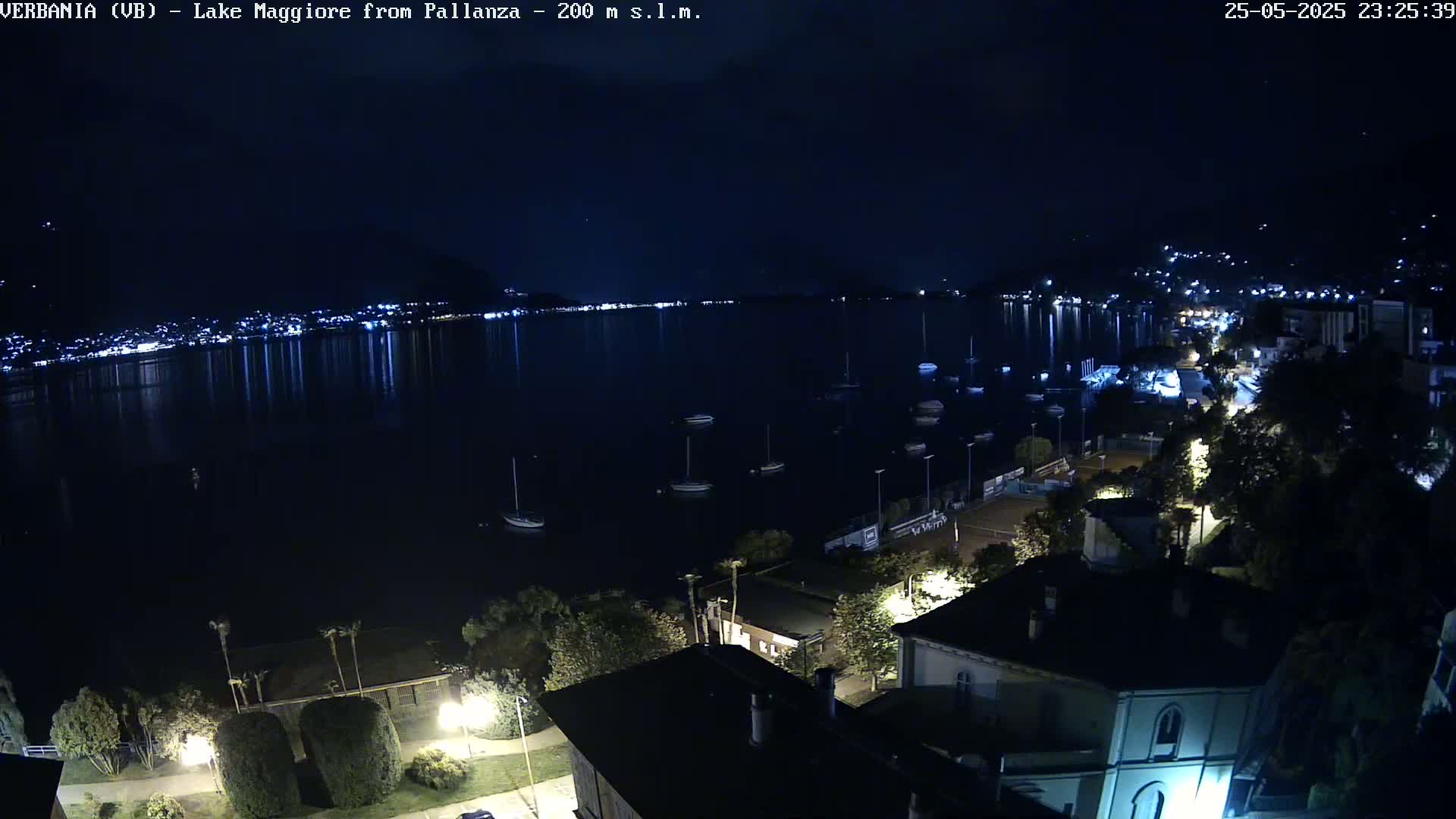 A nighttime view of a calm lake with boats moored, a shoreline town with lights, and dark hills in the background under a clear sky.
