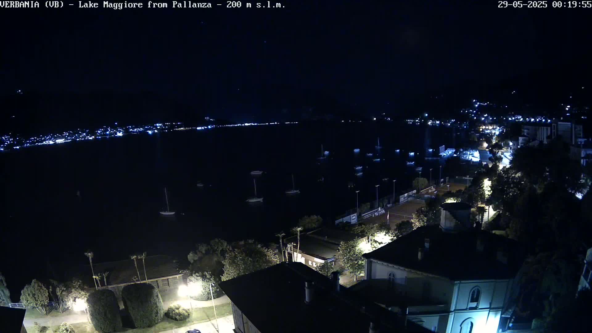 A nighttime, high-angle view of a dark body of water with several boats, a shoreline with buildings and lights, and a clear, dark sky.