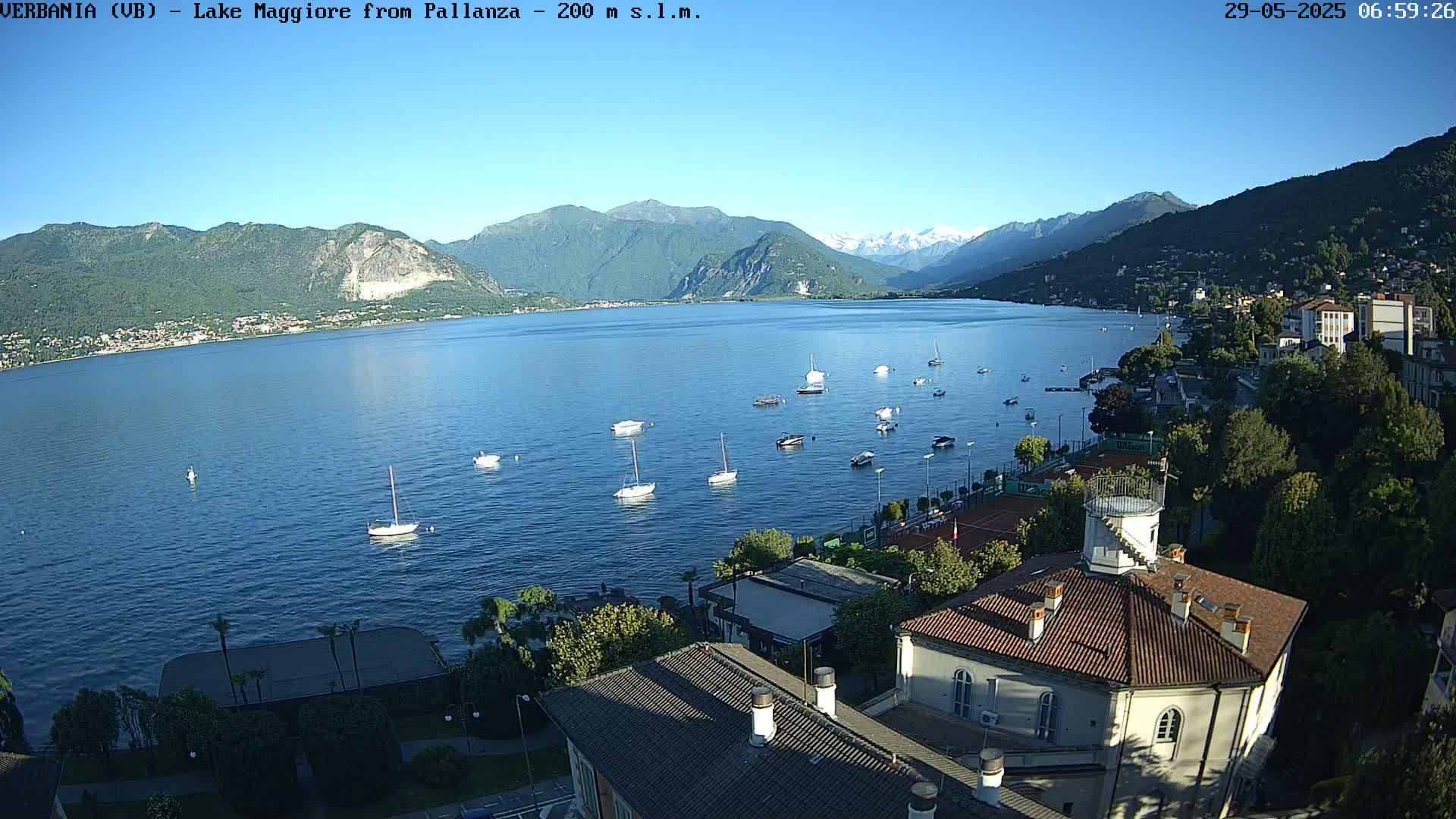 A calm lake with numerous sailboats is viewed from an elevated position overlooking a town nestled against mountains under a clear blue sky.