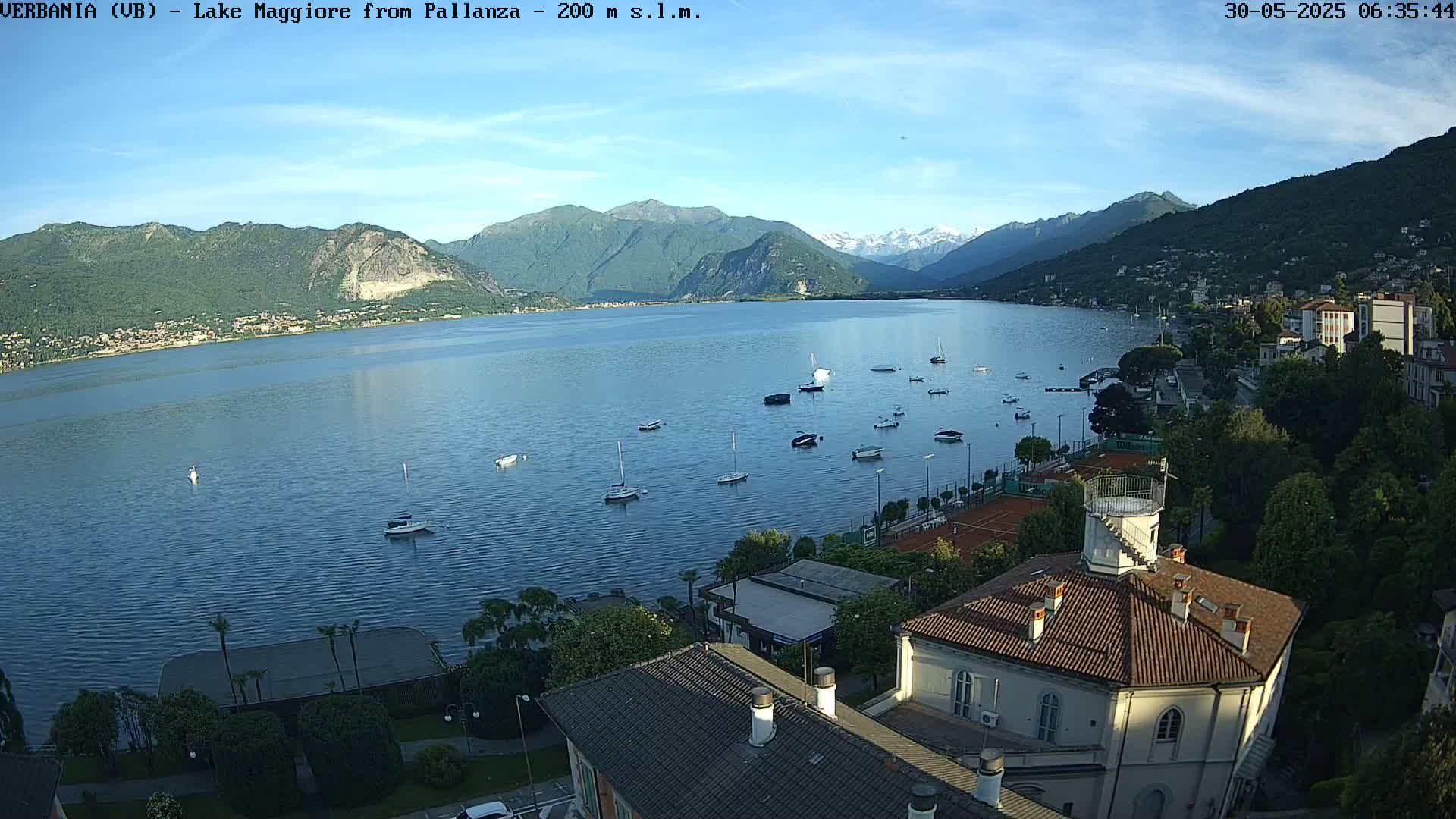 A calm lake with numerous boats is seen from an elevated vantage point, surrounded by lush green hills and mountains under a clear sky.