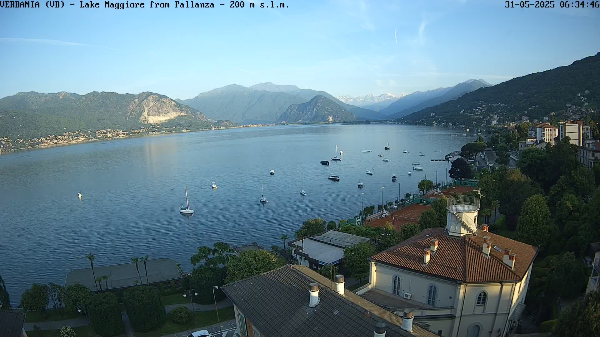 A calm lake with several boats is seen from an elevated vantage point overlooking a town nestled against mountains under a clear, bright sky.