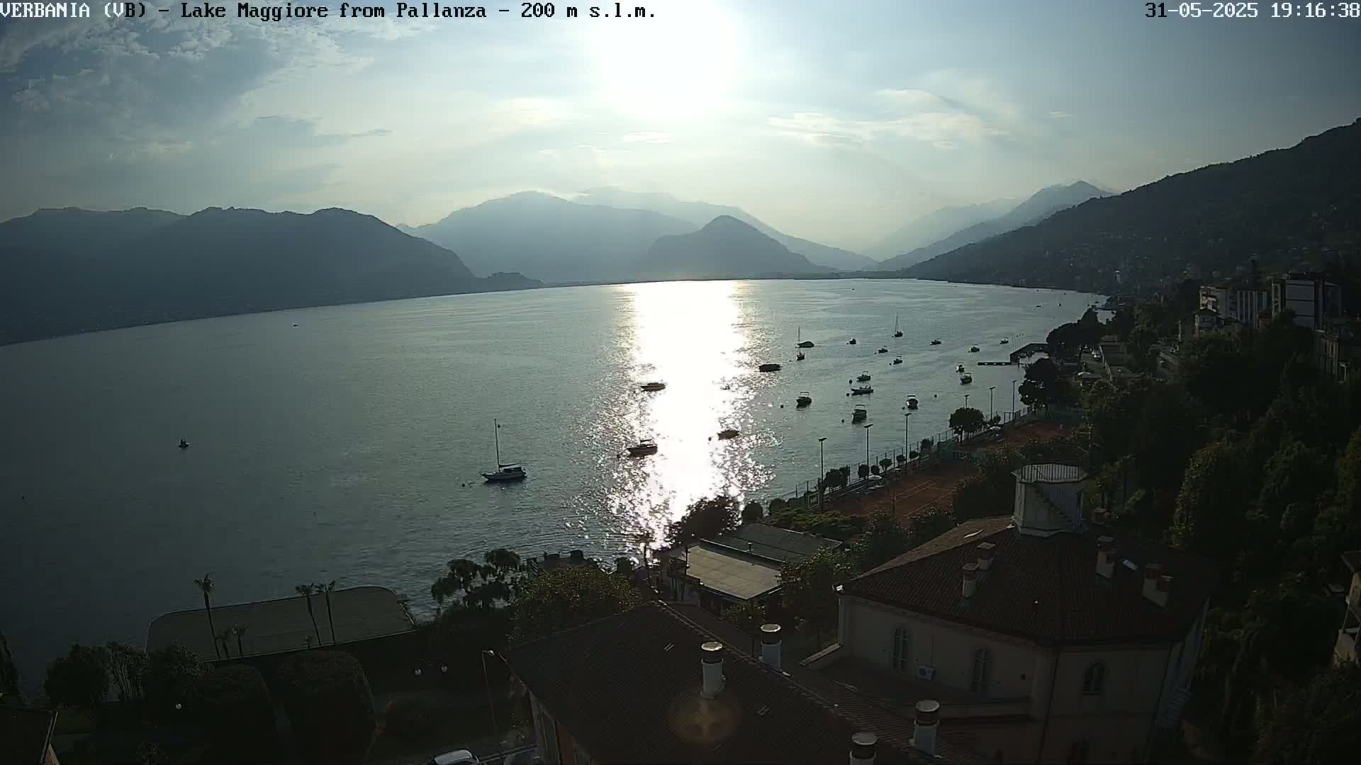 A calm lake reflecting the late afternoon sun, with boats scattered across its surface, is bordered by a hillside town with buildings and tennis courts under a partly cloudy sky.