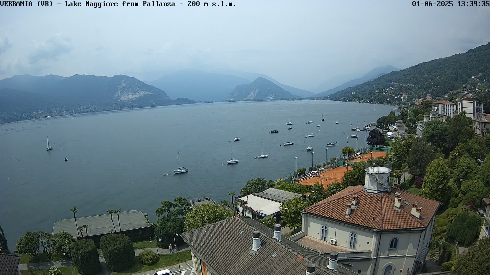A hazy view of Lake Maggiore from a hillside, showing several boats on the water, tennis courts, and buildings, with mountains in the background.