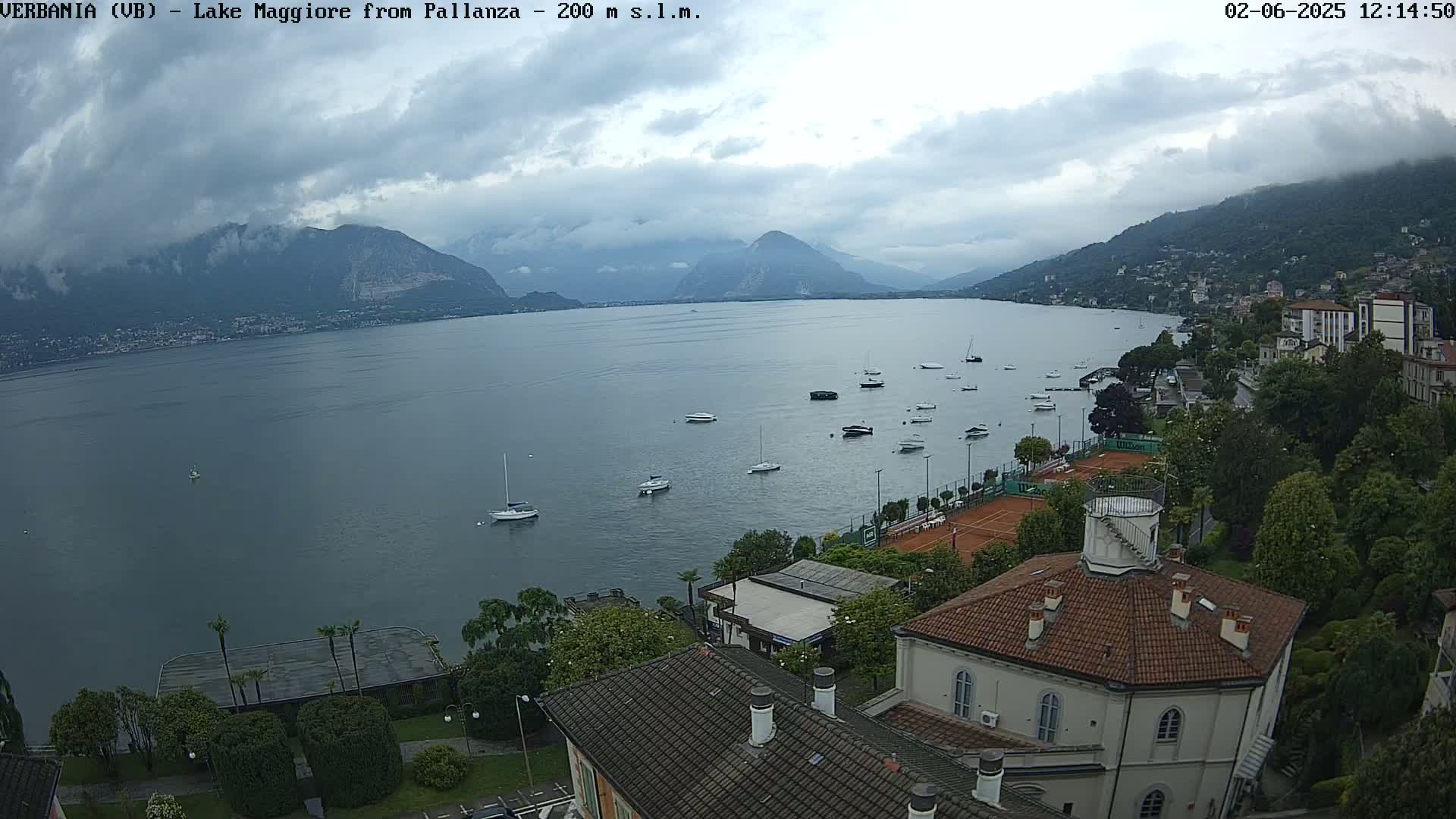 A calm lake with numerous small boats is nestled between mountains under an overcast sky; a town with several buildings, including a tennis court, is visible along the shoreline.