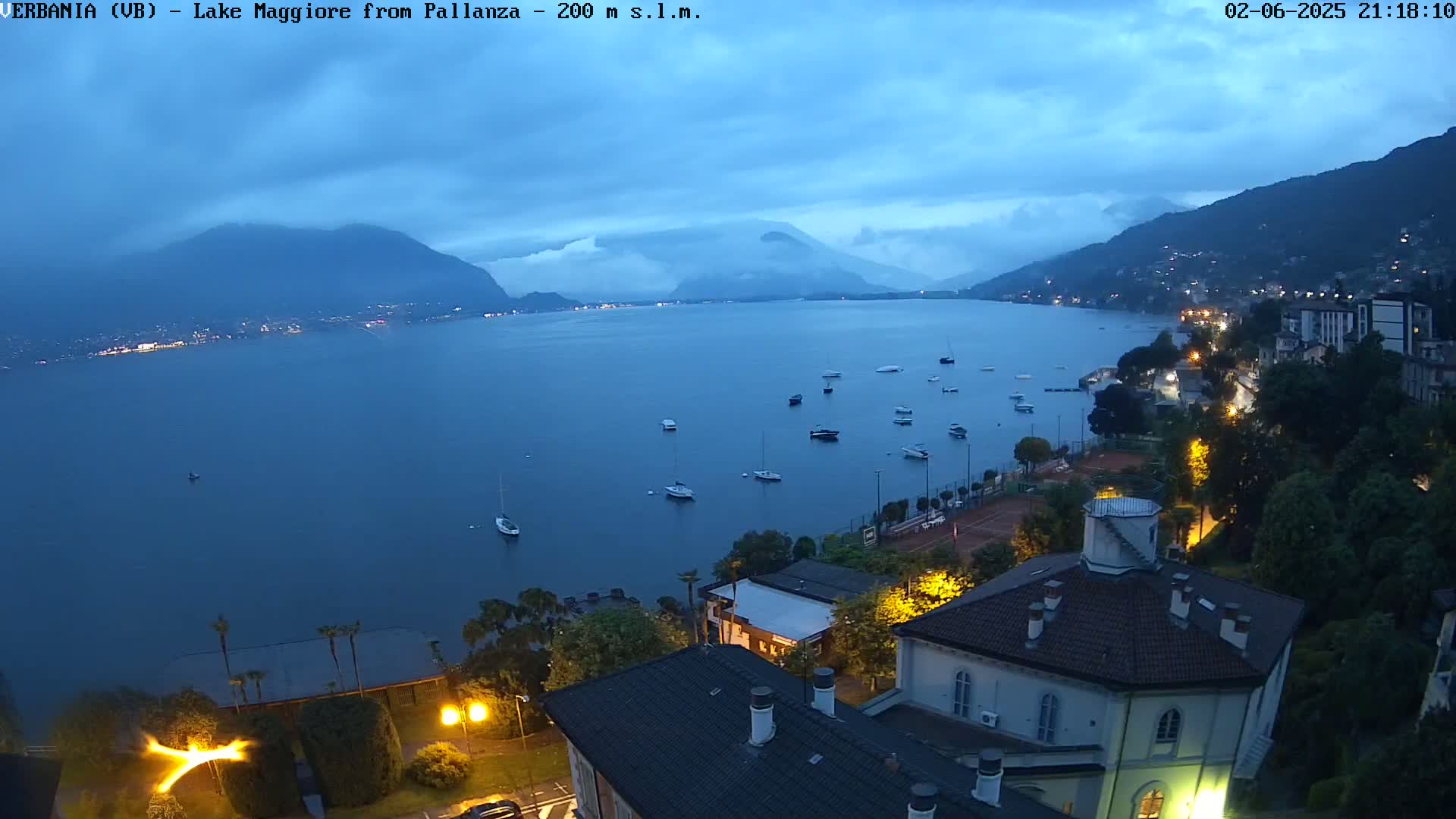 A twilight view of Lake Maggiore from a high vantage point, showing a calm lake with several boats, a shoreline with buildings and lights, and mountains in the background under a mostly cloudy sky.
