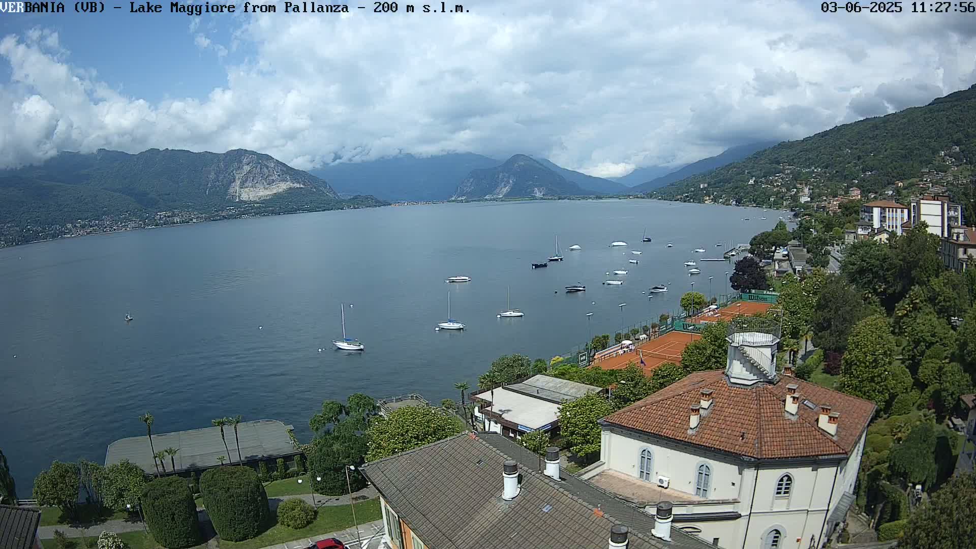 A partly cloudy day overlooking a lake with numerous sailboats, tennis courts, and buildings nestled among lush greenery against a backdrop of mountains.