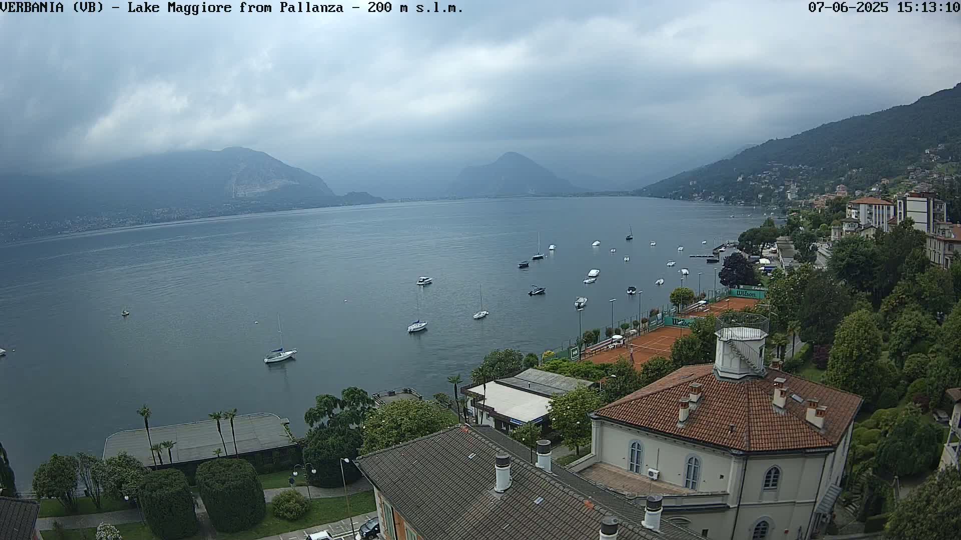 A view of a lake with several boats, a shoreline with buildings and tennis courts, and mountains in the background under an overcast sky.