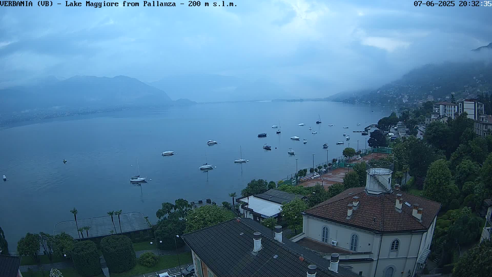 An overcast view of Lake Maggiore, with several boats moored on the water, overlooked by buildings and lush greenery.