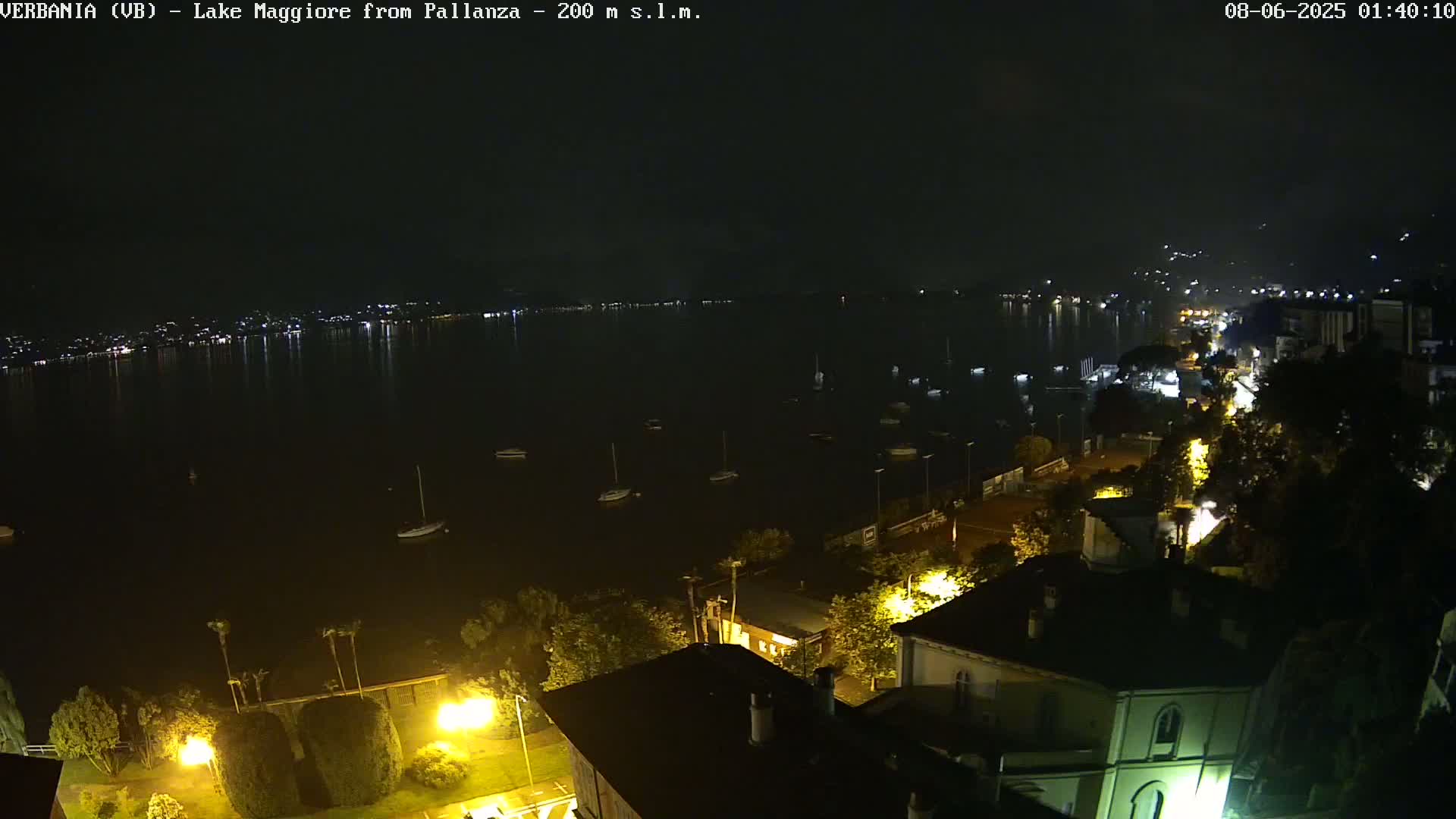 A nighttime view of a calm lake with several boats, a shoreline with buildings and streetlights, and a distant town illuminated across the water.