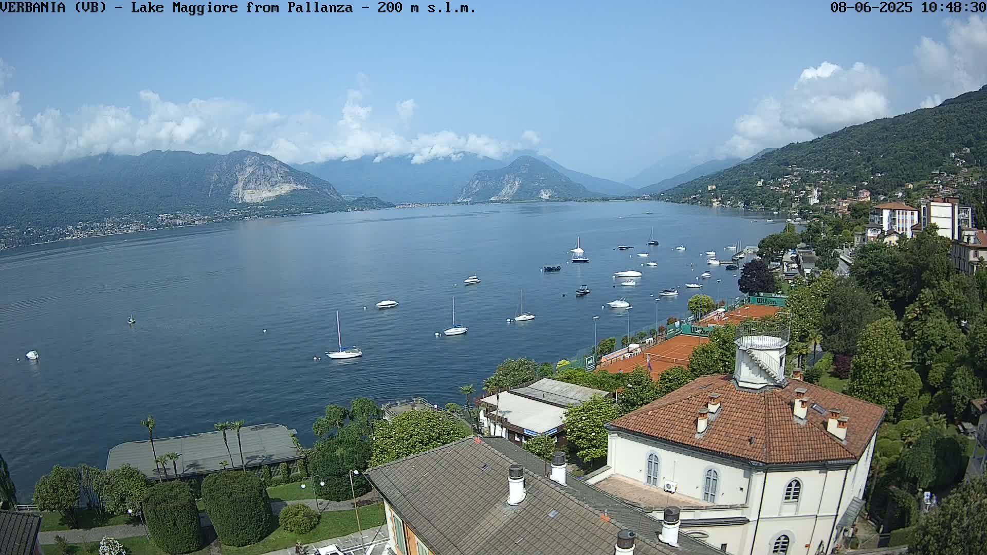 A sunny day overlooking Lake Maggiore, with numerous sailboats and motorboats scattered across the water, and a view of buildings and tennis courts along the shore.