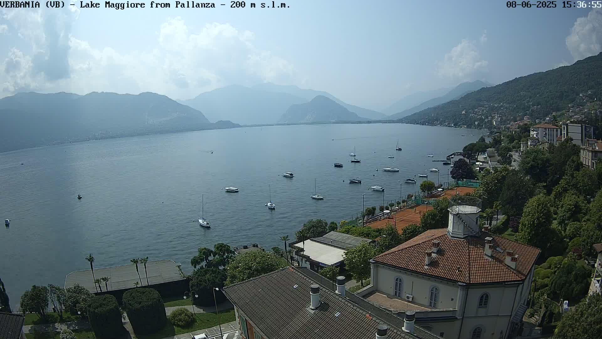 A lake with numerous boats is nestled between mountains under a partly cloudy sky, with a town and tennis courts visible along the shore.