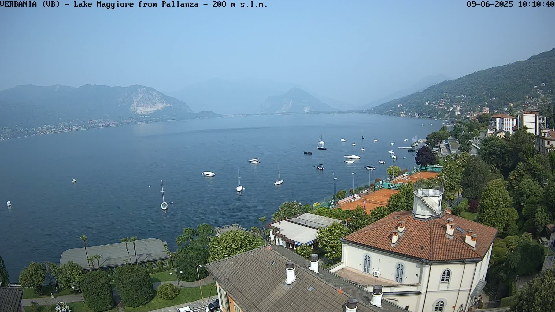 A hazy, sunny day overlooks a calm lake dotted with sailboats and motorboats, with buildings, trees, and tennis courts situated along the shoreline, backed by mountains.
