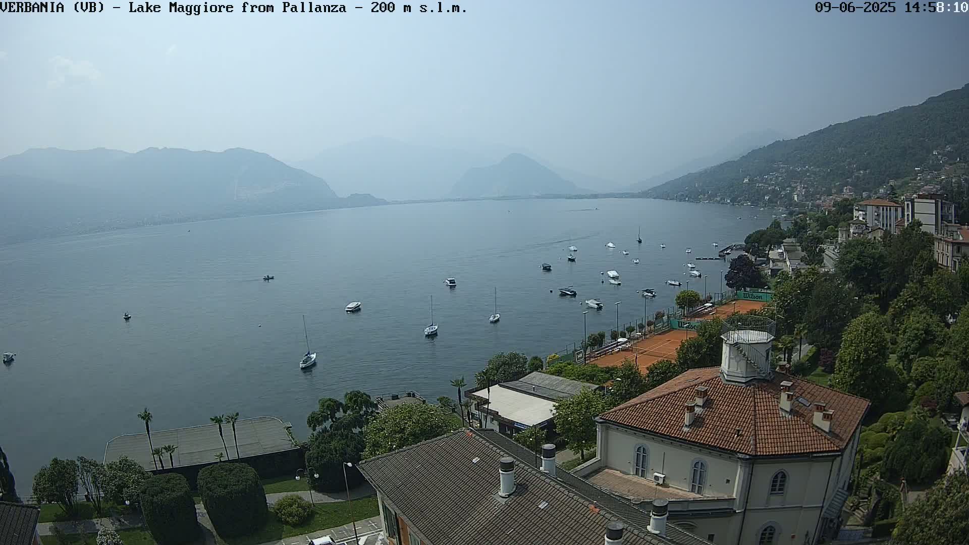 A hazy, sunny day overlooking Lake Maggiore, with numerous boats on the water and a tennis court and buildings visible along the shore.