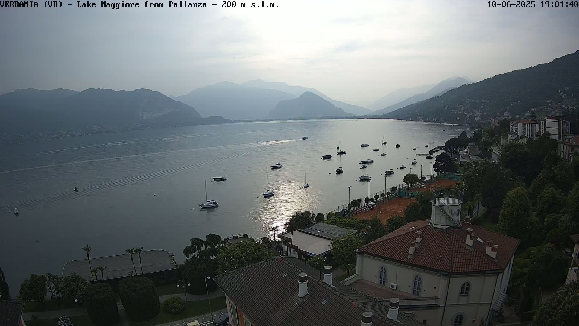 A calm lake with numerous boats is seen from an elevated viewpoint, overlooking a town nestled against hills under a hazy sky.